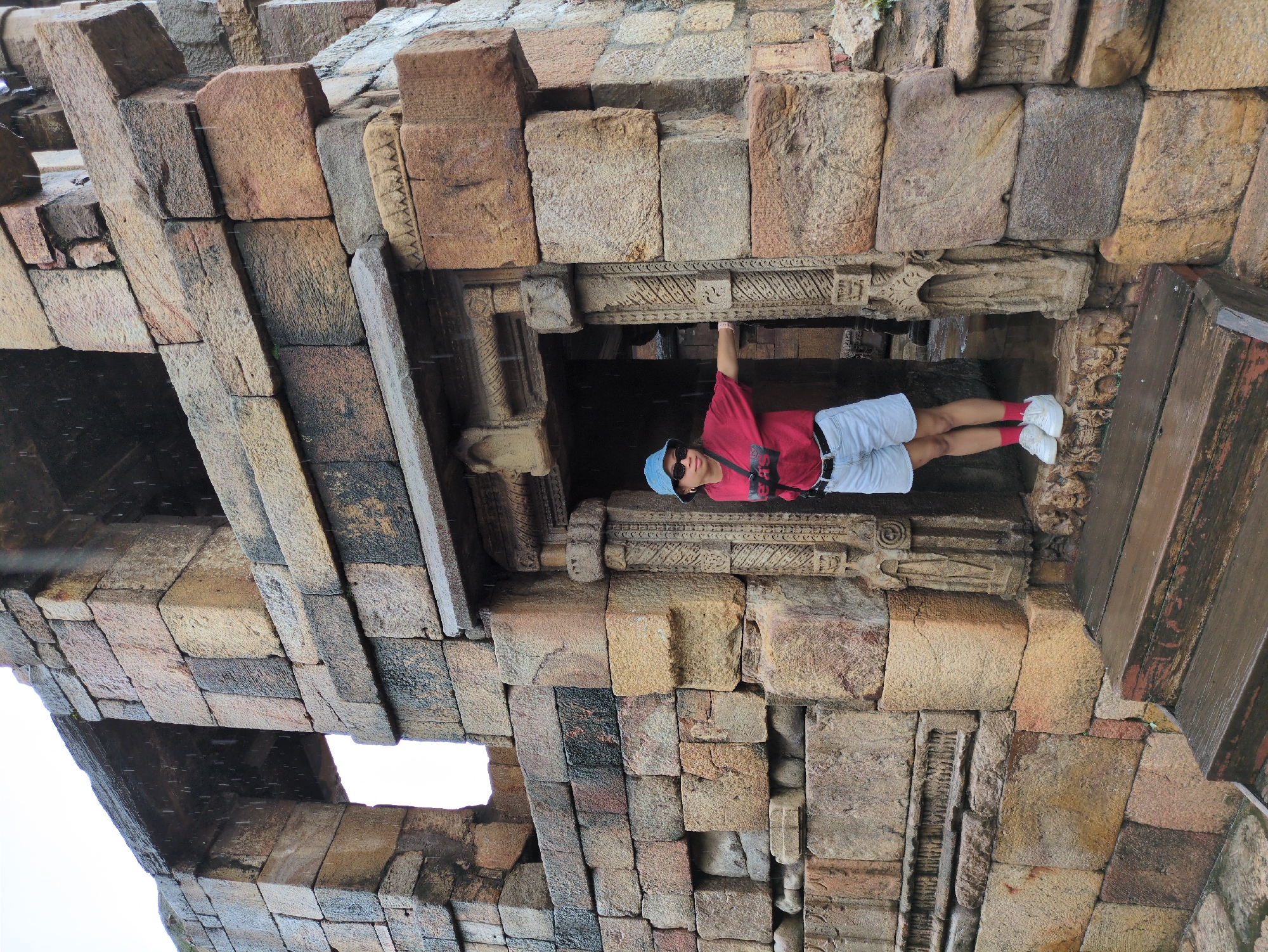 In Delhi, India, a person stands in the doorway of an ancient stone structure, possibly a temple or monument. The person, wearing a red t-shirt, light blue shorts, a blue hat, and white sneakers with pink socks, is looking towards the right. Their left arm is extended, resting on the stone frame of the doorway. The stone architecture is characterized by large, weathered blocks, some with intricate carvings. The area appears wet, with visible raindrops suggesting it is raining. The ground in front of the doorway is covered by wooden steps, also appearing damp. The background shows more of the stone structure, with openings that reveal a darker interior. The overall scene conveys a sense of historical significance and the presence of an ancient site experiencing inclement weather.