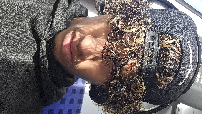 A male individual is depicted in a vertical close-up photograph, positioned from the chest upwards. The subject has medium-dark skin and wears a dark grey textured hoodie. Distinct curly, coily hair, primarily dark brown with prominent caramel-blonde highlights, is visible. A black baseball cap is worn backwards, with the adjustable snap-back mechanism visible. Black over-ear headphones are worn beneath the cap, partially visible around the ears. Faint facial hair, including a mustache and chin beard, is present. Direct sunlight creates intricate shadow patterns across the subject's face, particularly on the forehead, eyes, and nose, suggesting light filtering through a patterned window or overhead structure. The background is out of focus, revealing a blue patterned textile surface to the right, consistent with public transport seating. Indistinct bright light sources are visible to the upper left. The scene occurs within a form of public transportation, likely a train or bus, in Kamenz - Kamjenc, Germany.