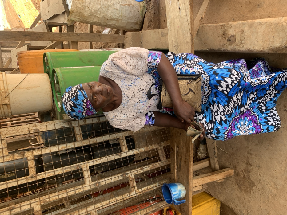 An adult female is seated on a light-colored wooden bench in a semi-enclosed outdoor area within Ube, Nigeria. She wears a vibrant blue, purple, white, and black patterned headwrap and a matching long skirt, with a white, lace-textured, short-sleeved top over an undergarment featuring the same pattern visible at her forearms. Her left hand holds a fanned stack of banknotes. A light brown, patterned shoulder bag rests on her lap. To her immediate left on the bench is a blue and white plastic mug. Behind her, a rusty metal grate structure with a padlock is positioned to the left, while several green plastic drums, an orange plastic jerrican, and a white plastic bucket are stacked to her right. A large, light-colored sack is visible further to the right. The ground surface is uneven earth. The woman's gaze is directed towards the right side of the frame.