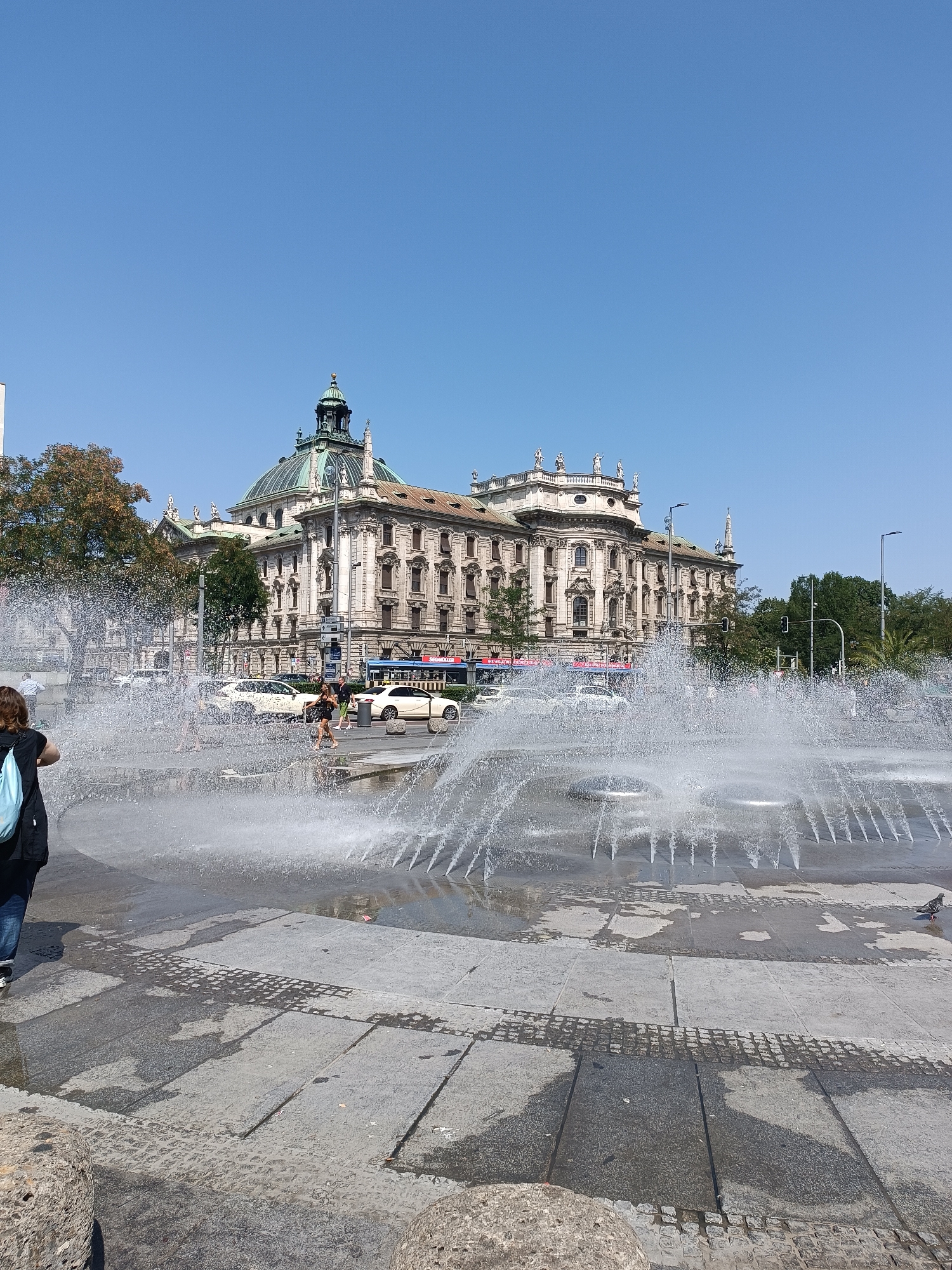 The image captures a daytime scene in Munich, Germany, likely Marienplatz or a similar central square, featuring a large fountain in the foreground with water spraying upwards. The water creates a misty effect, partially obscuring the background. Behind the fountain, a grand, ornate building with a distinctive domed roof and classical architectural elements is visible. This building is likely a significant landmark in Munich. Several people are present, some walking near the fountain and others in the background. Vehicles, including cars and what appears to be a double-decker bus, are on a street adjacent to the building, suggesting a public thoroughfare. Trees and a clear blue sky complete the urban landscape. The ground in the foreground is paved with stone slabs.