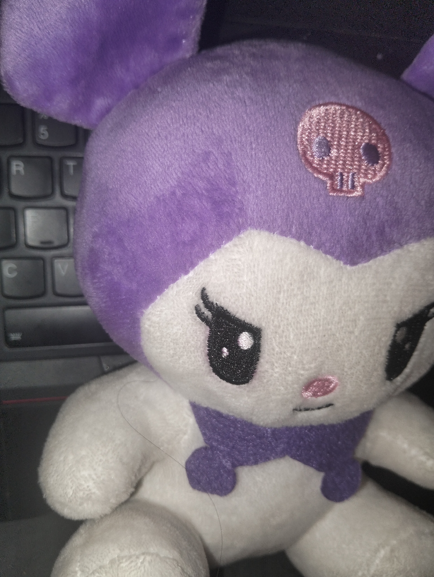 A close-up image of a Kuromi plush toy. The toy is primarily white with purple accents including its ears, part of its head, and a bow around its neck. A pink skull is embroidered on the purple part of its head. It has black eyes with white glints, and a pink nose. The toy is sitting on a black keyboard. The location in Dhaka, Bangladesh is unknown.