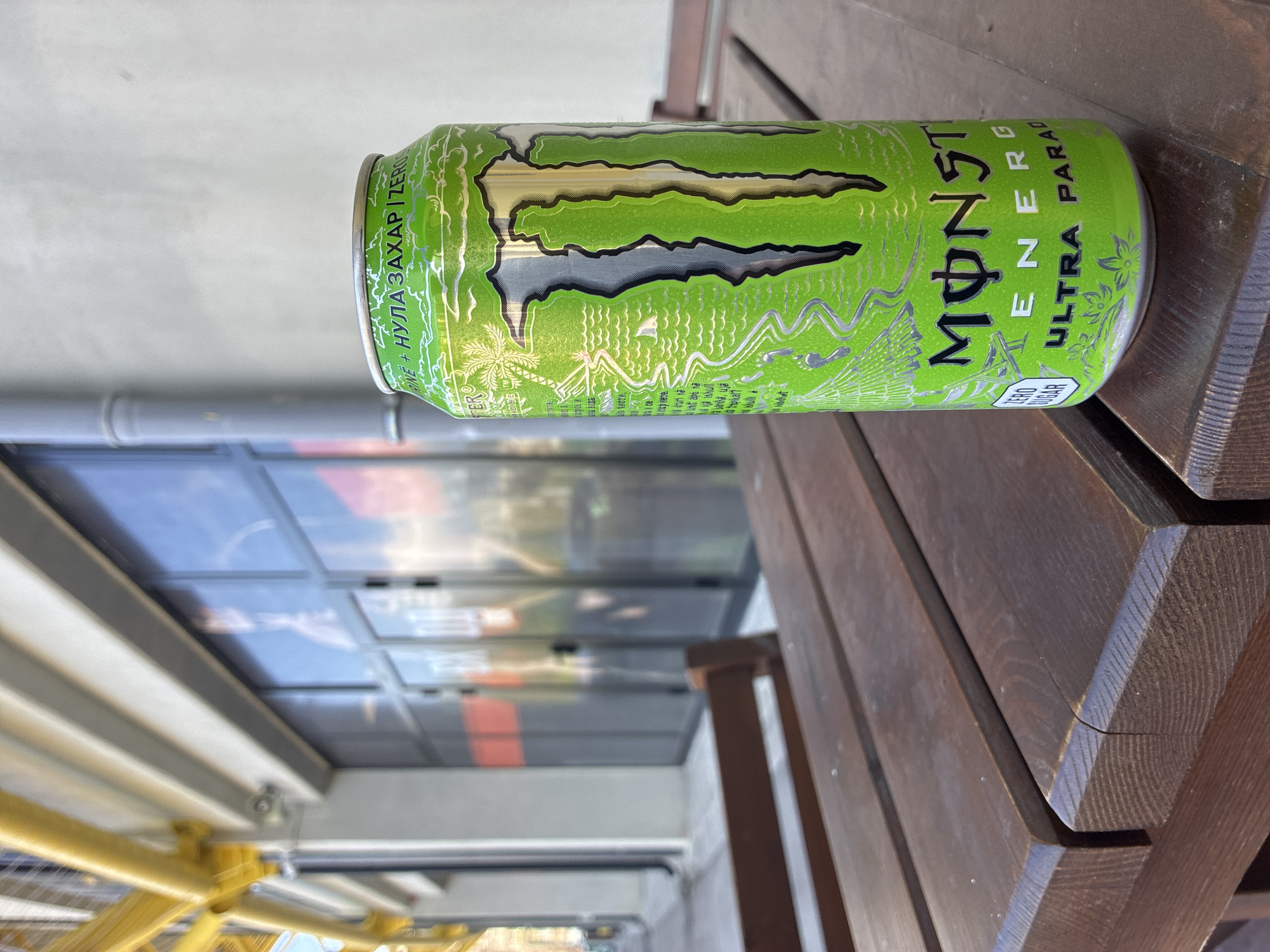An eye-level shot captures a vibrant green Monster Energy Ultra Paradise drink can resting horizontally on a dark wooden surface. The can features the prominent Monster logo in silver and black, with "ENERGY" and "ULTRA PARADISE" written in white. The design incorporates silver illustrations of palm trees and tropical waves, suggesting a refreshing, exotic theme. Below the main logo, "ZERO SUGAR" is visible, along with its Cyrillic translation "НУЛА ЗАХАР", indicating the product is sugar-free and sold in a Slavic-speaking region like Plovdiv, Bulgaria.
The wooden surface, likely a table or bench, exhibits distinct grain patterns and appears to be made of dark, sturdy wood. In the background, a blurred reflection in a large glass window or panel reveals elements of a modern building structure, including rectangular segments and hints of blue sky and clouds. A yellow metal frame and grey architectural components are also discernible in the soft-focus background, suggesting an outdoor or semi-outdoor setting such as a cafe terrace or balcony during daytime. No people are visible, and no specific activity is taking place beyond the can being set down. The bright lighting suggests it is daytime.