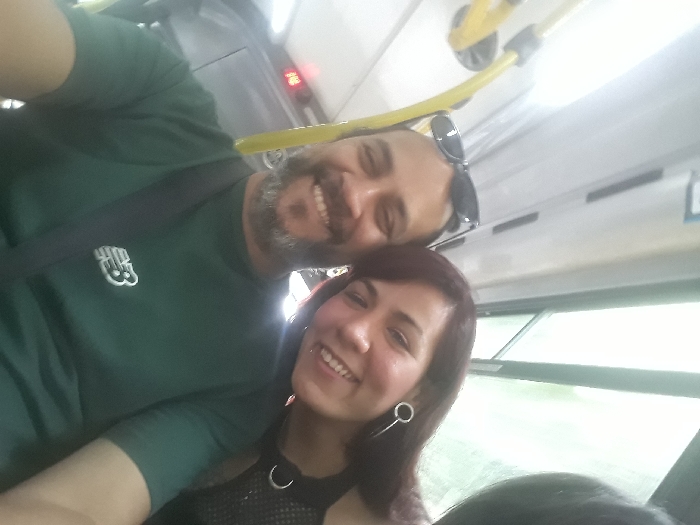 A selfie captures a man and a woman smiling inside a bus. The man, in a green shirt with a logo, has a gray beard and sunglasses atop his head. The woman, with dark hair and hoop earrings, wears a mesh top with a pendant. Yellow handrails are visible above, along with a window showing greenery outside. A seatbelt crosses the man's chest. The overall scene conveys a casual and happy moment during transportation.