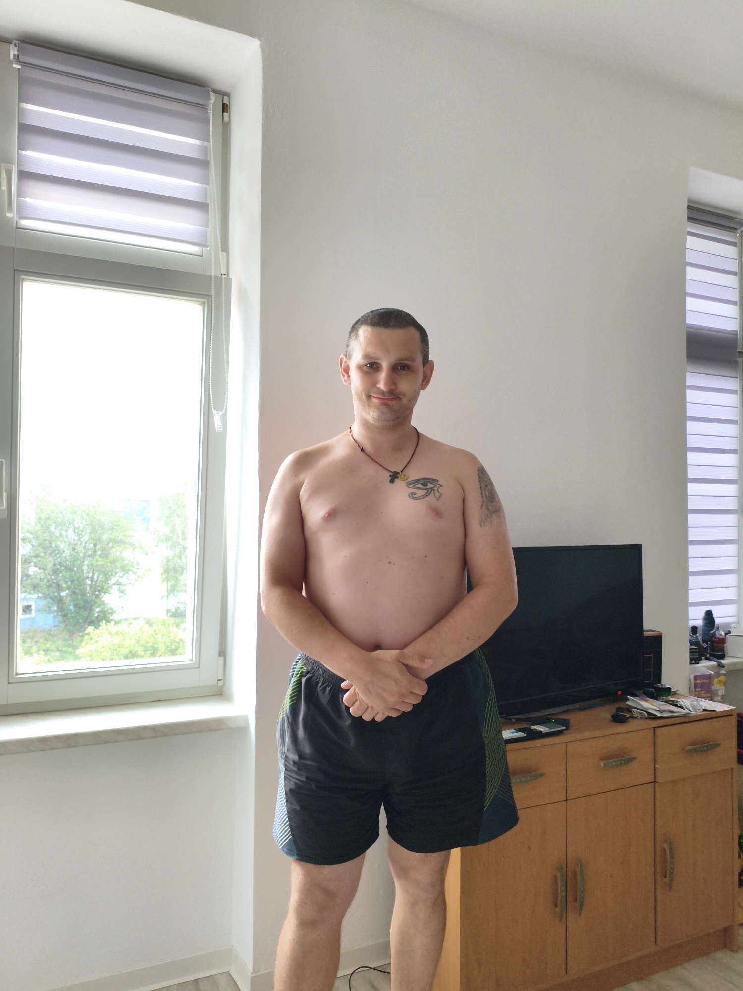 A shirtless man stands in front of a window and a cabinet in an indoor setting. The man is of Caucasian ethnicity, with short dark hair, and is wearing dark athletic shorts. He has a tattoo of an eye on his chest and another on his left arm. His hands are clasped in front of him. To his left is a large window with horizontal blinds, revealing greenery and a building outside. To his right, a wooden cabinet with drawers and doors is visible, topped with a television and various items, including bottles and papers. The walls are white, and the floor appears to be light-colored wood. The scene is indoors, possibly a room in a residence. The city of Wolgast, Germany, is not depicted or referenced in the image.