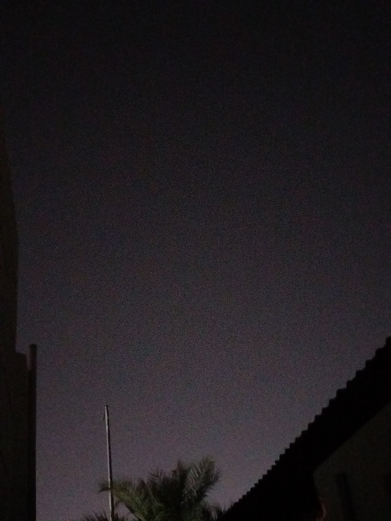 This is a nighttime photograph taken in Al Mas'oudi, United Arab Emirates. The image is largely dark, with a textured, grainy appearance. The sky is a dark, overcast gray. In the foreground, the dark silhouette of a roofline is visible on the right side of the frame, angled upwards. To the left of the roofline, a tall, thin, light-colored pole or antenna extends vertically into the sky. Below and to the left of the pole, the dark green fronds of a palm tree are visible. On the far left of the image, a dark vertical structure, possibly a building or wall, is partially visible. The scene appears to be an outdoor urban or suburban setting at night.