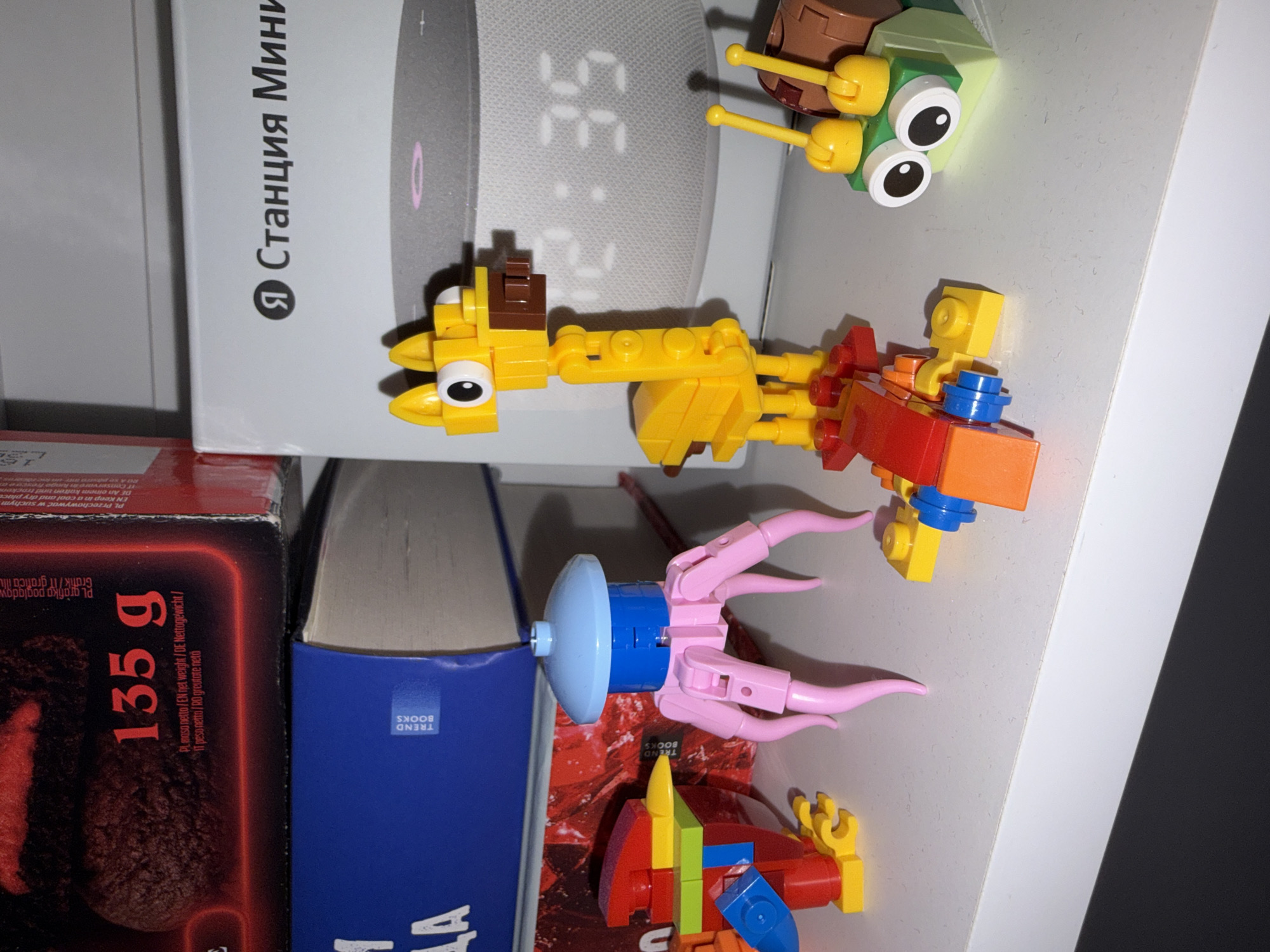An indoor scene, likely a shelf in a home in Perm, Russia, displays a collection of objects including a smart speaker, books, and several colorful Lego creations.
In the upper left, a white box for a "Я Станция Мини" (Yandex Station Mini) smart speaker is visible, with the speaker's display showing "2:35". Perched on top of this speaker box is a light green and brown Lego creature with prominent googly eyes and two yellow drumstick-like pieces. Directly in front of the smart speaker, a larger yellow Lego creature, possibly a dragon or monster, features big eyes and a vibrant body composed of red, orange, and blue bricks.
Below these, nestled between a blue book bearing the "TREND BOOKS" logo and a red box, is a playful pink and light blue Lego figure, resembling an octopus or jellyfish with multiple tentacles. Another multi-colored Lego creation, predominantly red, yellow, and blue, is visible further down, in front of the red box. The red box appears to be food packaging, indicating a "135 g" net weight and providing text in multiple languages, including "PL masa netto / EN net weight", alongside storage instructions.
The scene is lit by artificial indoor light, and the arrangement of items suggests a personal, lived-in space. No human activity is depicted.