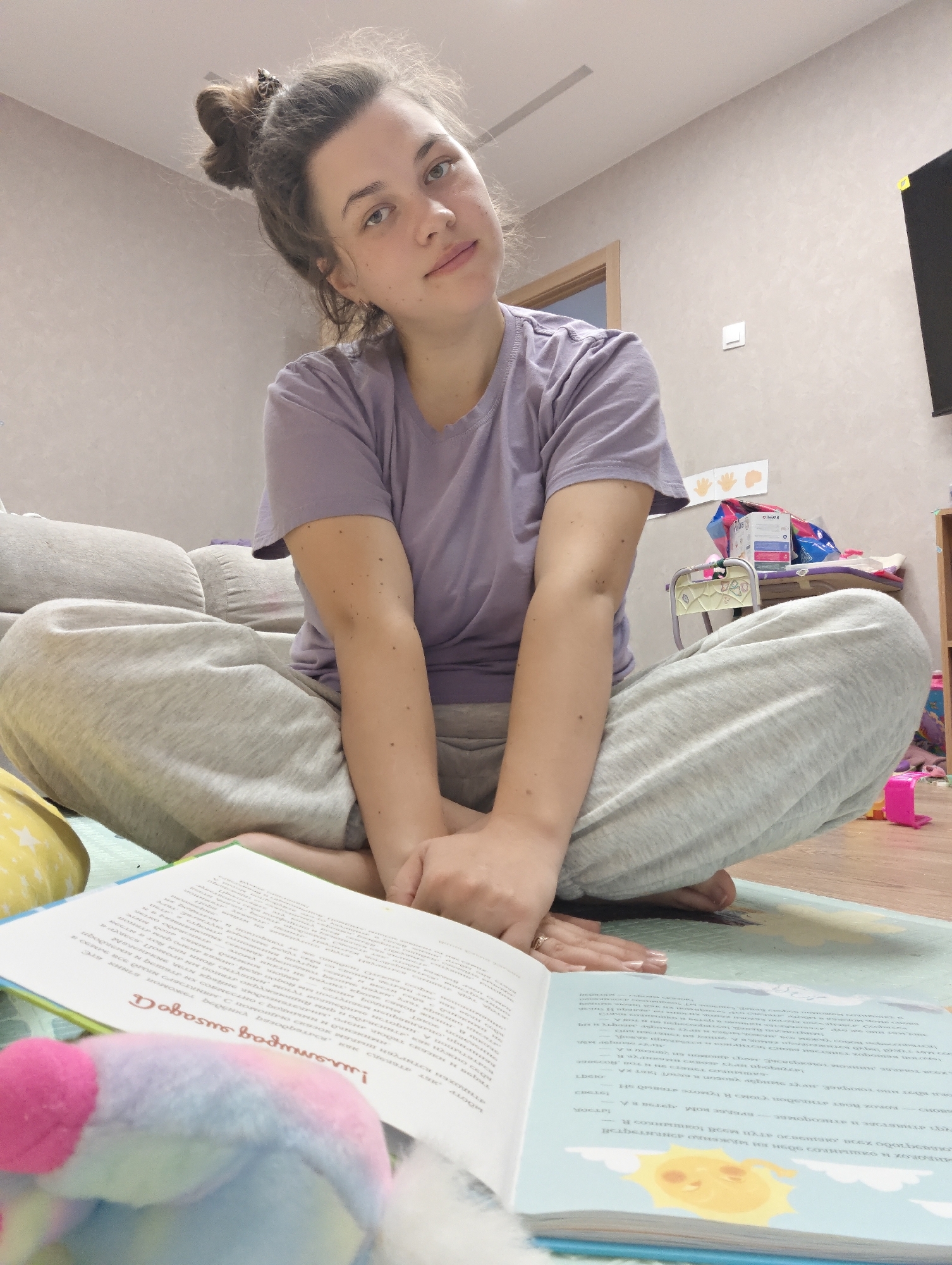 The image depicts an indoor residential scene in the city of Chaikovsky, Russia. A female subject, appearing to be an adult with light brown hair styled in a messy bun, is seated cross-legged on a light blue playmat. She is wearing a light purple short-sleeved t-shirt and light grey sweatpants. Her bare feet are tucked beneath her legs, and her hands are clasped together, resting in front of her, with a gold-colored ring visible on the ring finger of her left hand. She looks directly at the camera with a neutral to slightly smiling expression, revealing light-colored eyes and minor freckling on her face and arms. An open book, displaying Cyrillic text and an illustration of a yellow sun with a face on the right page, lies on the mat before her. A blurred, multi-colored plush toy is partially visible in the bottom left foreground.
In the background, a light grey upholstered couch is situated to the left. The wall behind the subject is a textured light beige. A wooden doorway with a light frame is visible in the mid-left background. To the right of the subject, a white light switch is mounted on the wall. Below the switch, three white paper handprints are taped to the wall. Further right, a small light-colored child's chair, a stack of boxes including one marked "OLLOWE," and various colorful toys are visible on the floor and within a wooden shelving unit. A dark, rectangular screen is mounted high on the far-right wall, displaying a small yellow marker on its top right corner. The room is uniformly lit, suggesting ambient or overhead lighting.