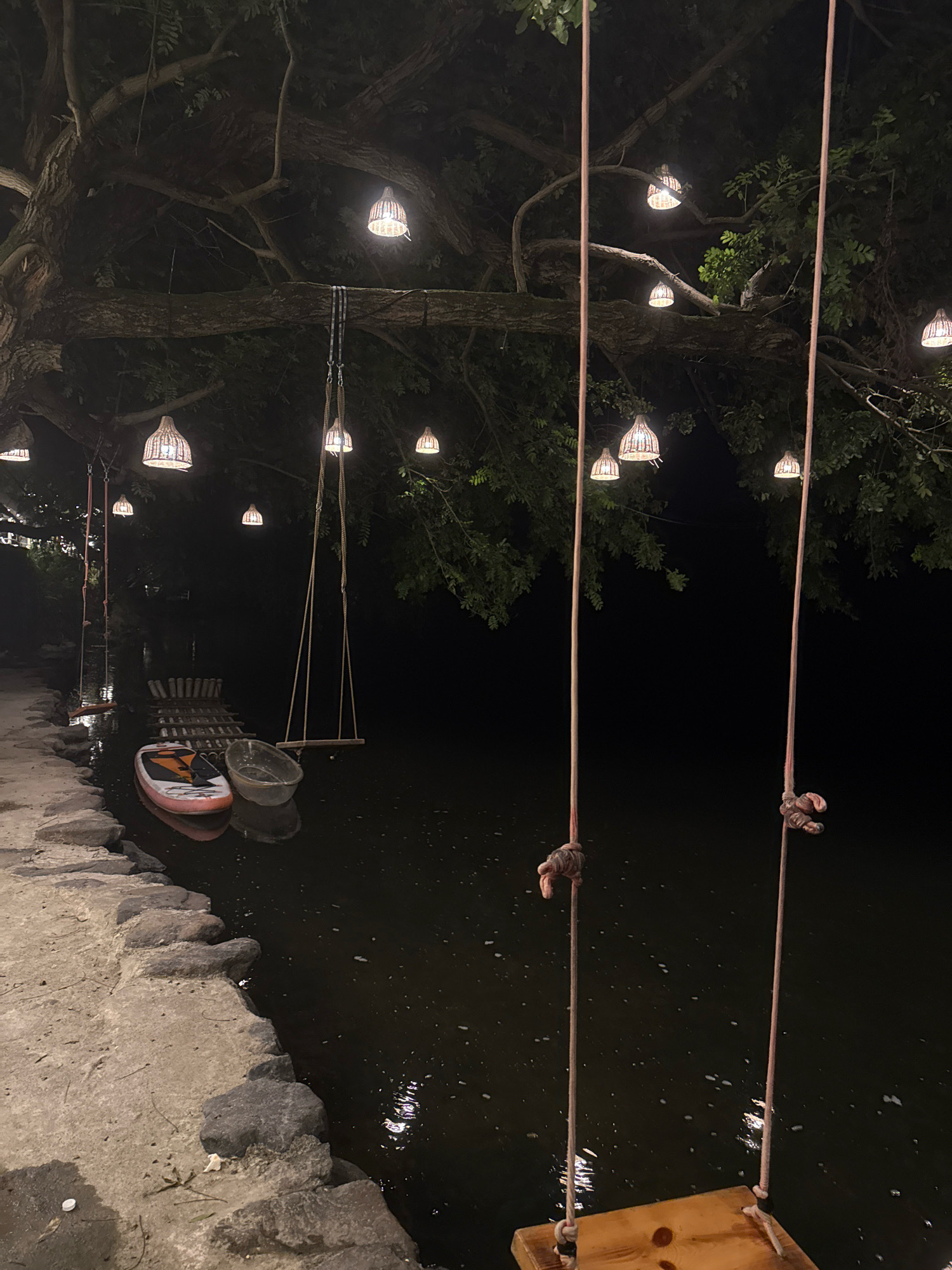 The image depicts an outdoor nighttime scene in Yiwu, China, likely a waterfront area. Large trees with branches overhead are adorned with numerous hanging lights featuring woven lampshades. Two swings hang from the tree branches over a dark body of water. In the foreground, a stone pathway lines the water's edge. Further out on the water, a paddleboard and a small boat or vessel are visible. The water reflects the lights from the trees, creating a serene ambiance.