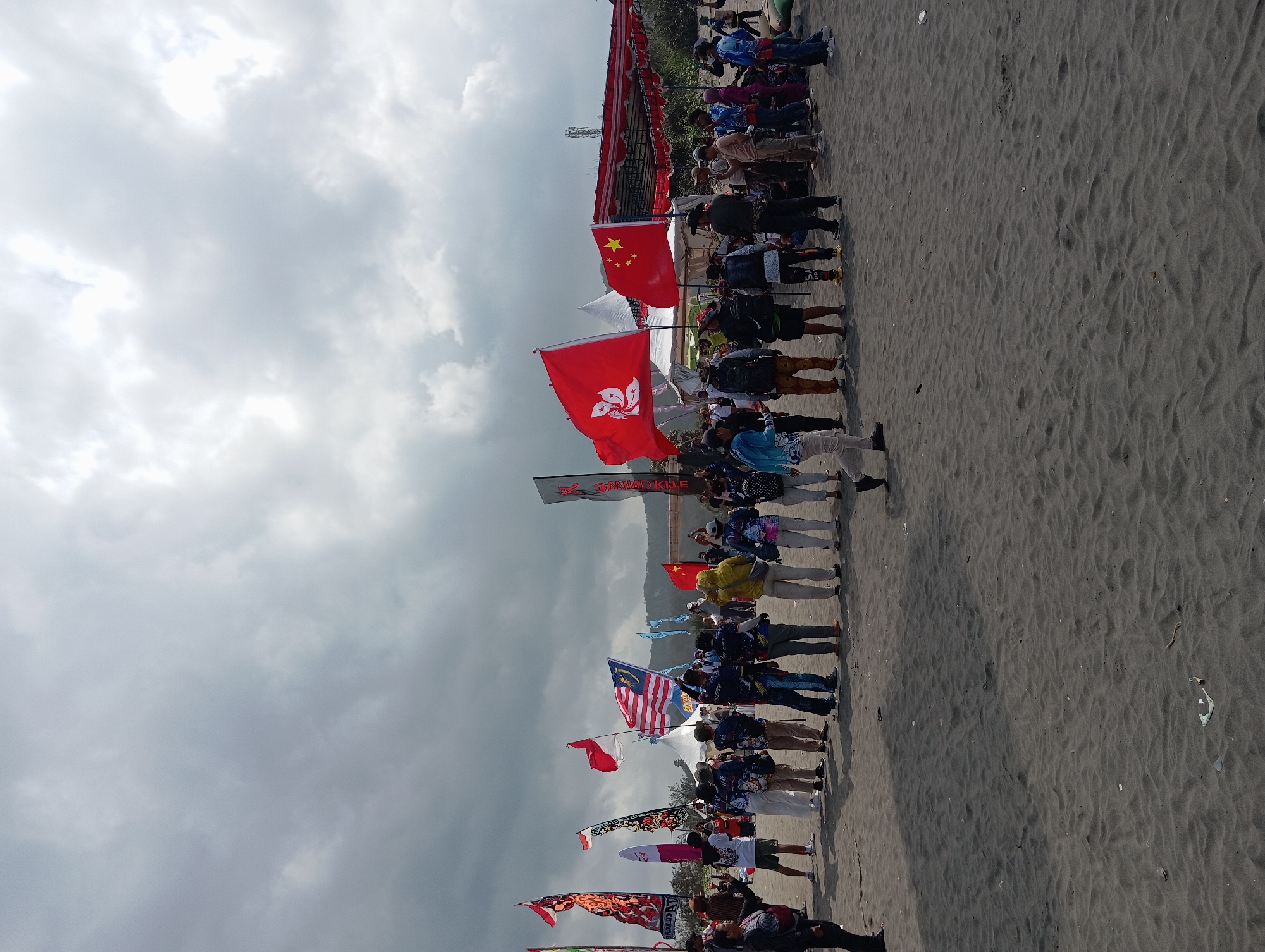 The image depicts a beach scene in Parangtritis, Indonesia, under a cloudy sky. A group of people, mostly adults, are gathered on a dark sand beach. Many are wearing matching shirts, suggesting they are part of an organized event or team. Several flags are prominently displayed, including the flag of Malaysia, the flag of China, and a red flag with a white flower emblem, possibly representing Hong Kong. Other flags with various designs are also visible.
In the background, there are tents and a covered structure, hinting at a gathering or festival. The sand is dark and textured with ripples. Some individuals are holding up their phones, possibly taking pictures or videos of the event. The overall atmosphere appears to be one of a public gathering or outdoor activity on the beach.