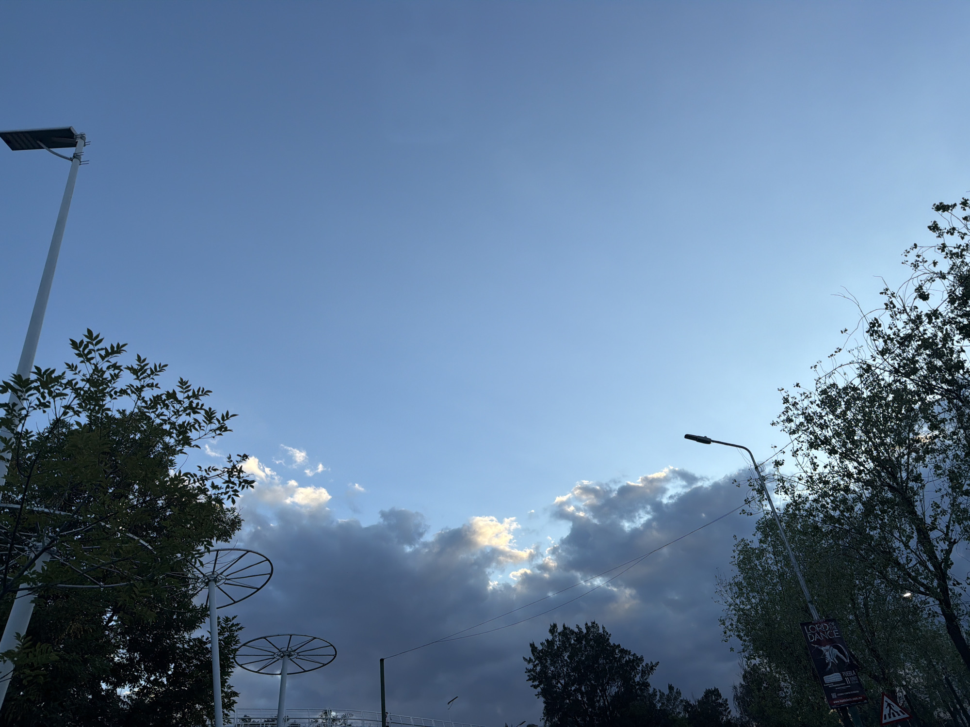 The image depicts a cityscape with a blue sky partly covered by clouds. Trees are visible on both sides of the frame. A tall, white lamppost stands on the left, and a standard street lamp is on the right. Metal structures with circular elements appear between the trees on the left. A sign and pedestrian crossing sign are visible on the right.
