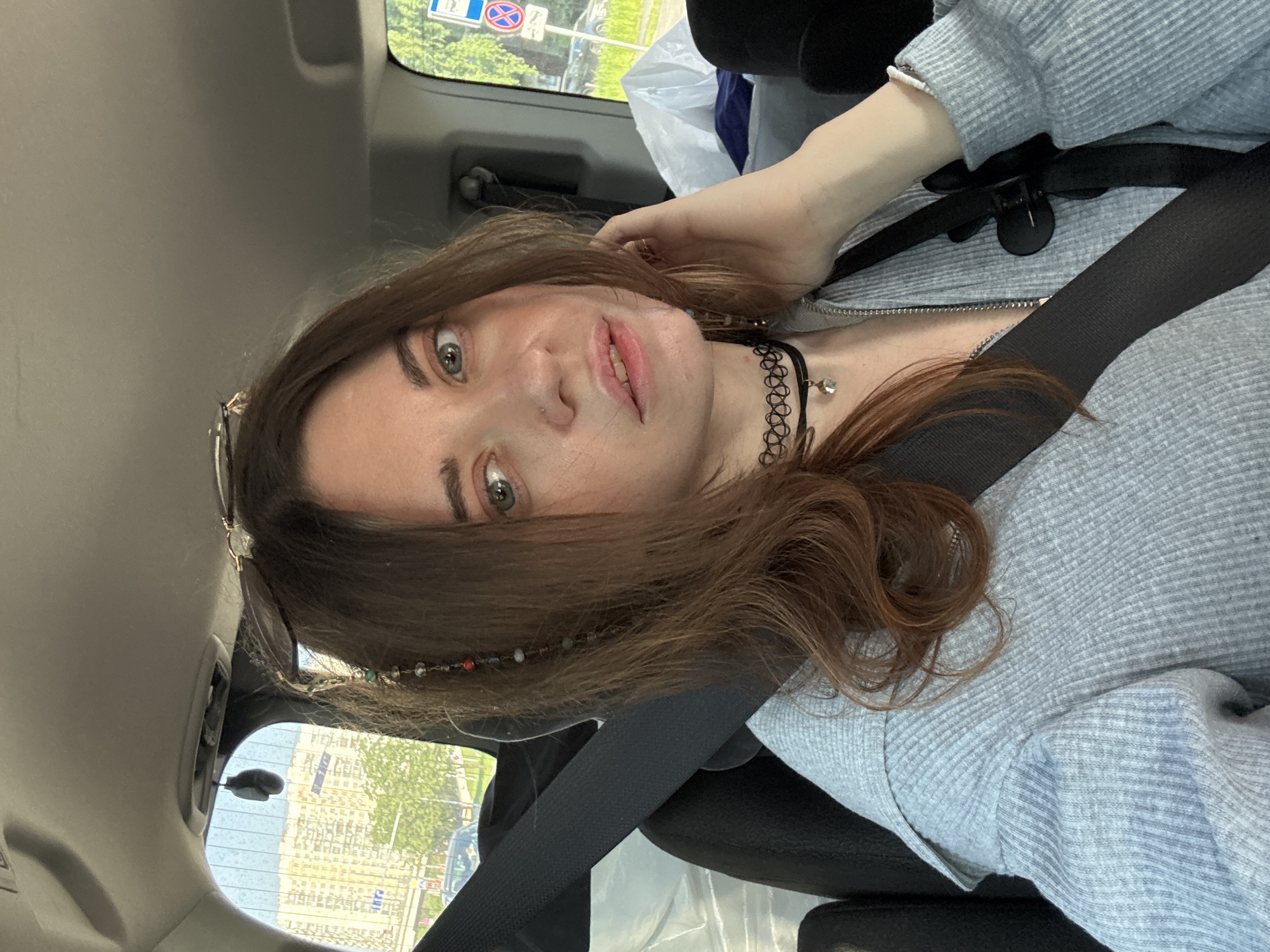 A woman is sitting in the front seat of a car, wearing a grey jacket and a black choker necklace. A seatbelt is fastened across her body. Her hair is brown and wavy, with a beaded accessory near her forehead. Outside the car window, there is greenery and buildings, with a road sign visible. The location is Moscow, Russia, as indicated by the road sign.
