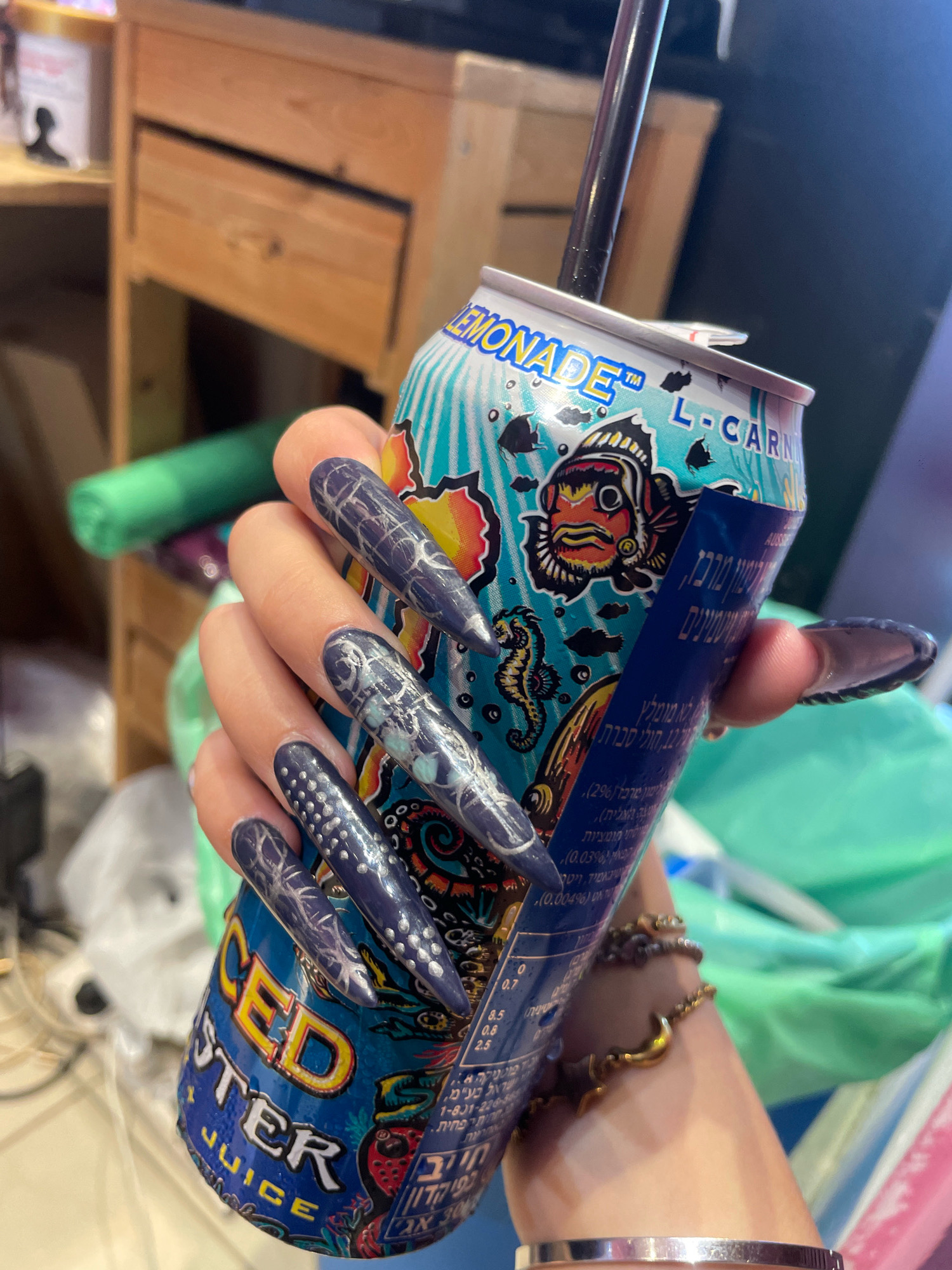 A person with long, pointed, dark blue nails adorned with intricate silver designs is holding a can of Monster Energy Juice. The can features a blue and turquoise design with various sea creatures and bold lettering. A black straw extends from the top of the can. The background appears to be an indoor setting, possibly a store or a room, with wooden furniture and some green and white bags visible. There are no explicit indicators of the city of Raanana, Israel, in the image.
