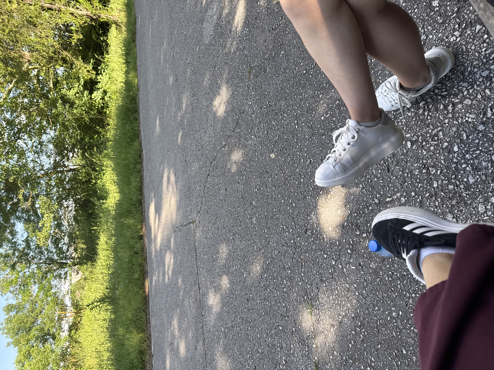 The image shows a section of asphalt road in Elhovo, Bulgaria, bordered by a grassy area with trees. Three pairs of legs and shoes are visible: one pair with white sneakers, one with white platform sneakers, and another with black Adidas sneakers and a water bottle next to the right foot. The perspective is directly above the legs, looking downward onto the road. Sunlight is present, casting shadows.