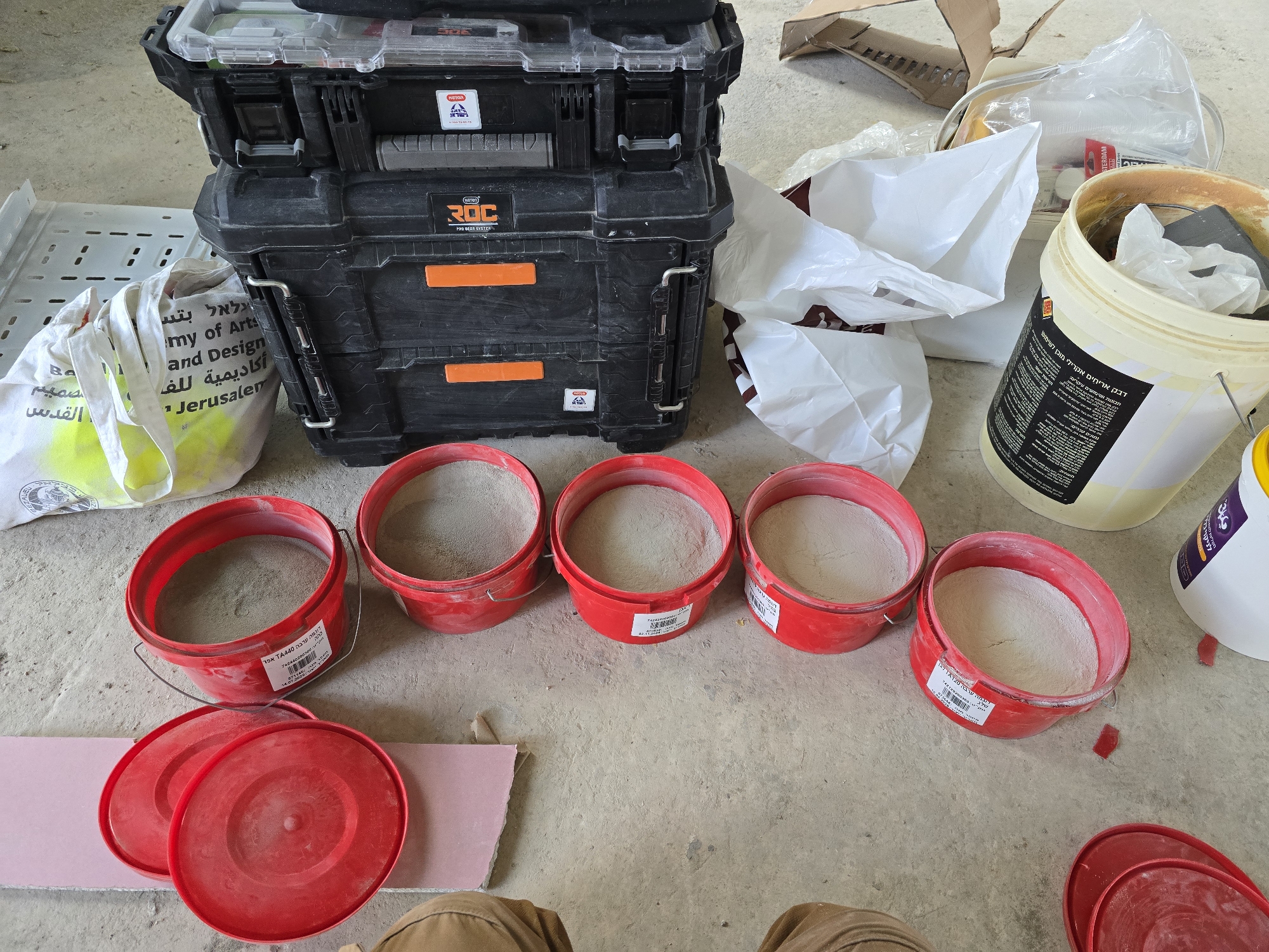 A ground-level shot reveals a clutter of construction-related objects against a light-grey concrete floor. The main items include red plastic buckets filled with a fine, light-grey powder, likely plaster, and a stack of black toolboxes, the second one labeled "ROC PRO GEAR SYSTEM" in orange. Additional elements include various white plastic bags, a light-yellow bucket with a partially visible label in Hebrew, and a partially visible bag labeled "Jerusalem Academy of Art and Design." The scene suggests an indoor setting, possibly a workshop or renovation site. A small, torn piece of drywall lies on the floor, along with the knee of a person wearing khaki pants.