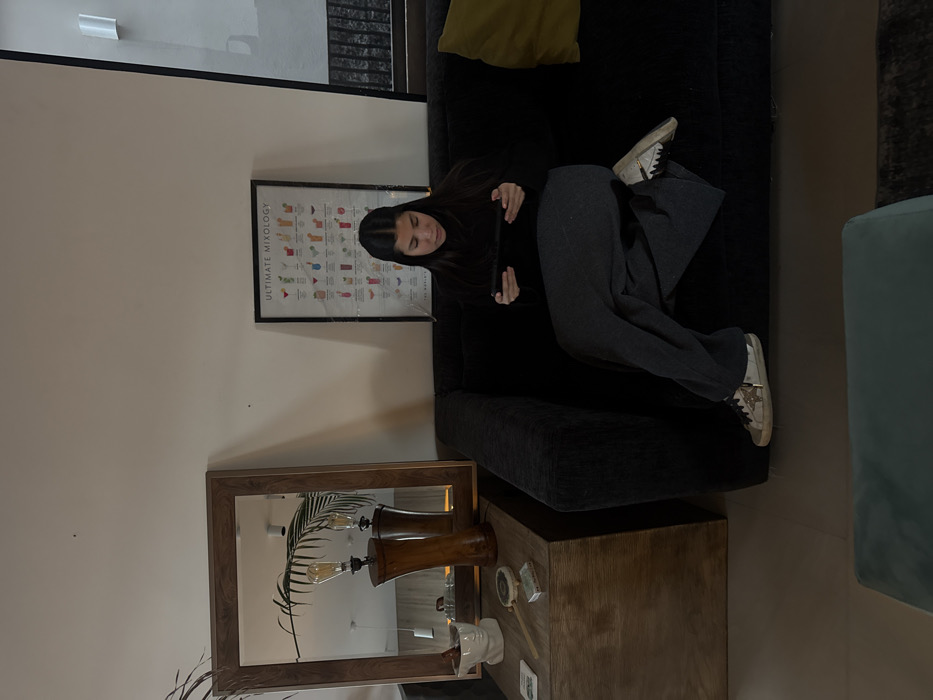 A young woman is sitting on a dark couch, looking at a smartphone. She wears black clothing and grey pants, with white sneakers on her feet. Behind her is a framed poster displaying beverage recipes. In front of her, there's a wooden cabinet with a mirror displaying a lamp. The picture is taken indoors, with a modern decor.