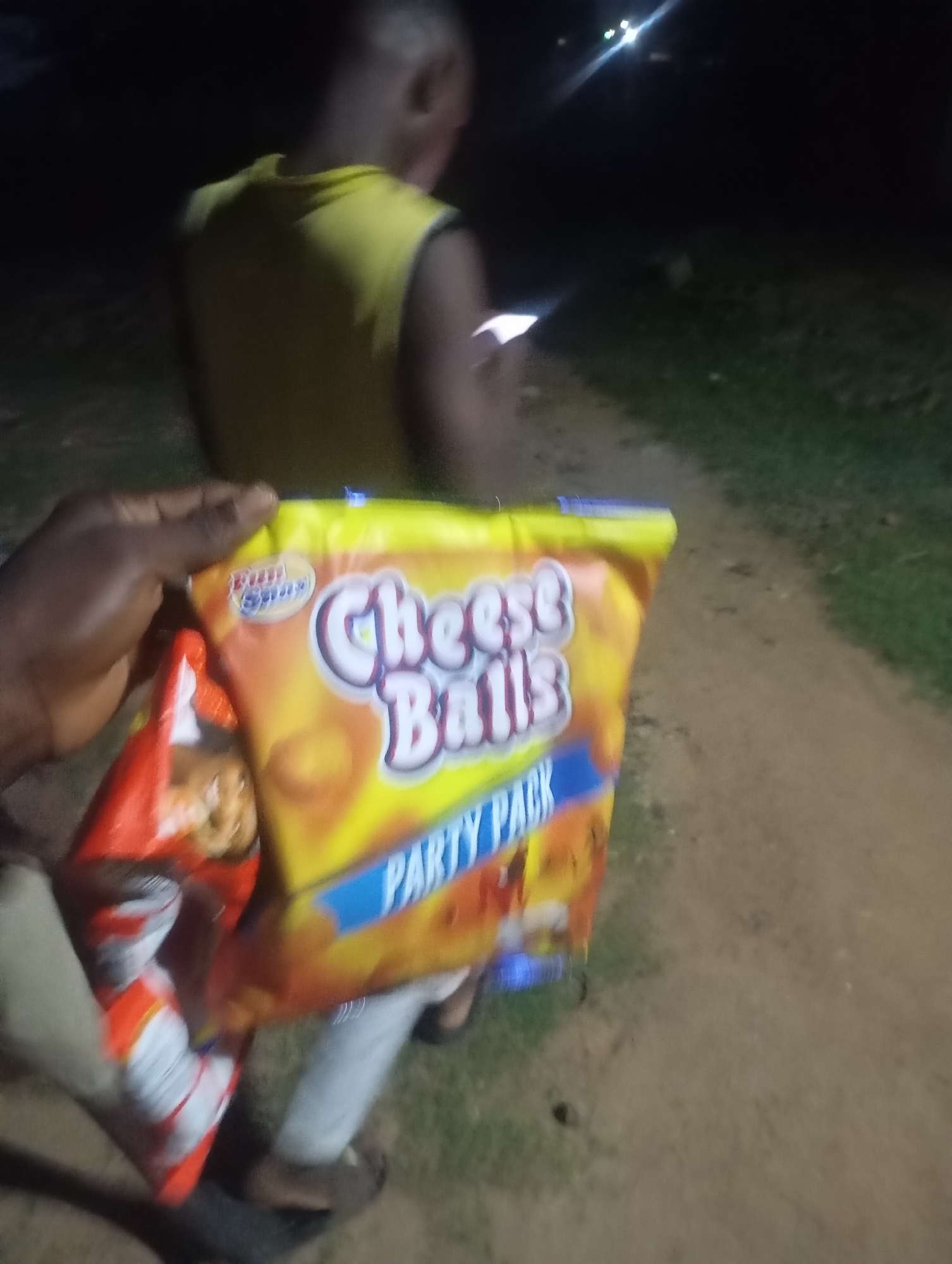 The image depicts an outdoor, dimly lit nocturnal scene in the city of Unknown, Unknown. In the lower-left foreground, a dark-skinned left hand holds two snack packages. The more prominent package is a large, brightly colored yellow and orange bag of "Cheese Balls PARTY PACK" from the "Bun Smax" brand. Partially obscured beneath it is a red and white snack bag, which appears to contain cookies or crackers. In the mid-ground, a second individual is heavily blurred and viewed from the back, appearing to walk away from the camera. This individual has closely cropped hair and wears a bright yellow sleeveless top. Their right hand is holding an illuminated device, possibly a smartphone, casting a subtle glow. The ground beneath the individuals consists of a light brown, possibly sandy or dirt path on the left, transitioning to a darker green, grassy area on the right. In the distant background, several distinct points of artificial light, exhibiting lens flare, are visible against the dark sky, suggesting distant infrastructure or lighting. There is no direct interaction evident between the two visible individuals; the foreground subject appears to be presenting the snacks to an unseen recipient or the photographer.