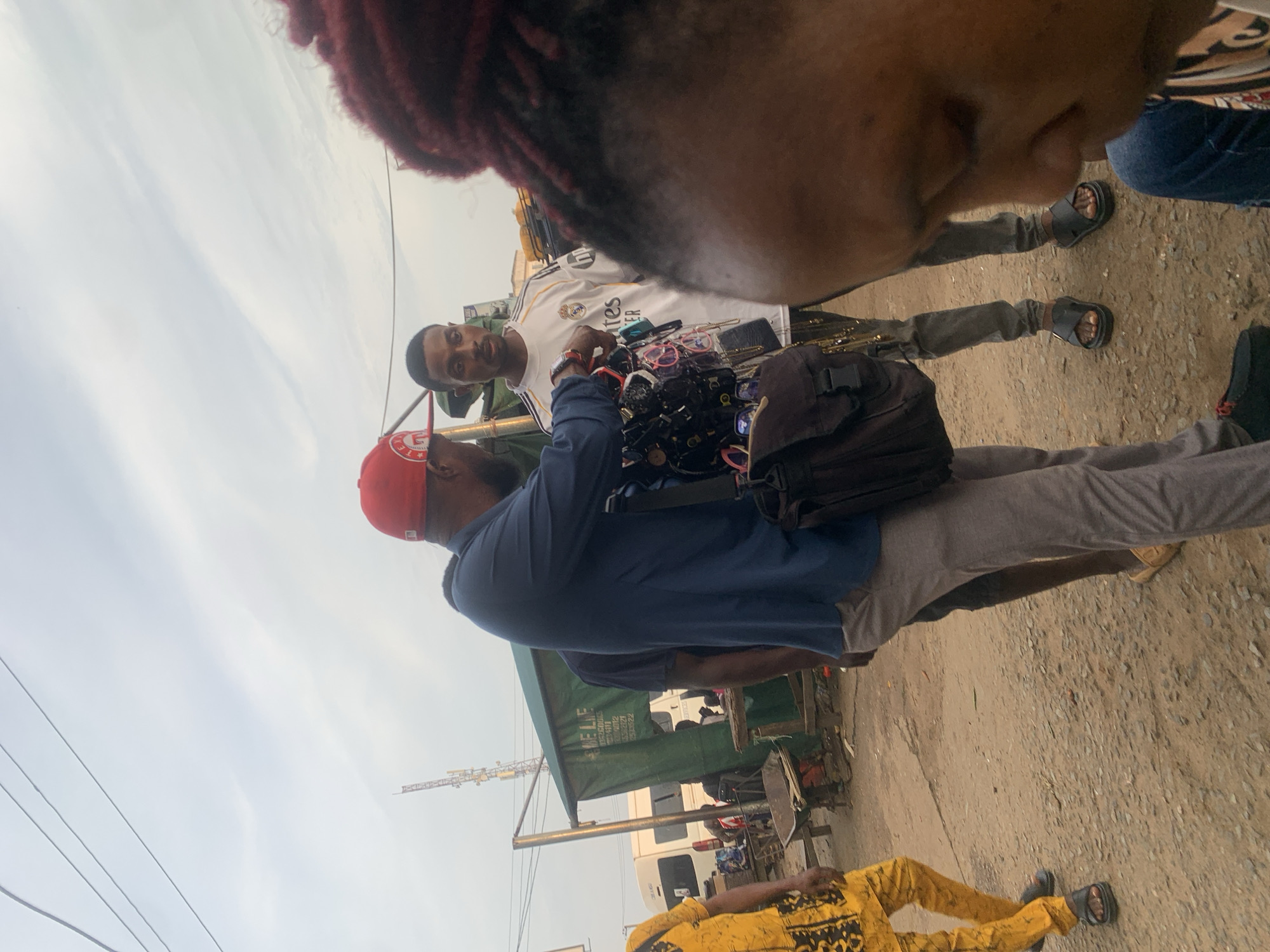 The image shows a street scene in Elelenwo, Nigeria, with several individuals interacting in what appears to be a market environment. In the foreground, a person's head and shoulder are visible, facing away from the camera. To the left, a person wearing a yellow and black patterned outfit and black sandals is walking away. In the center, a man in a navy blue shirt, gray pants, and a red cap is showing a display of sunglasses to another man who is wearing a white Real Madrid jersey, gray pants, and black sandals. The man in the jersey is looking at the sunglasses. Behind them, a green tent or stall is set up, with text visible on it, and a white van is parked nearby. The ground is unpaved and appears to be dirt and gravel. A telecommunications tower is visible in the background against a cloudy sky. The overall scene suggests a bustling marketplace with vendors and customers.
