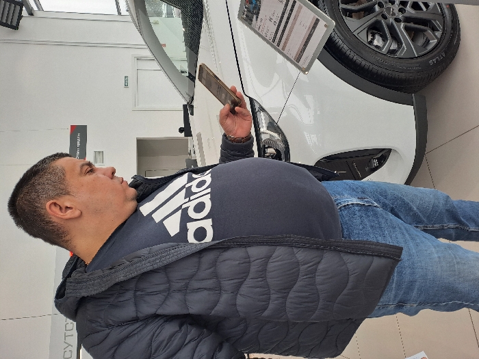 A man in a navy blue Adidas T-shirt and a quilted black jacket stands beside a white car, holding a smartphone. He is looking at a price display attached to the car. He is wearing blue jeans. The car has black rims and the word "ATLAS" is visible on the tire. The photo appears to be taken in a car dealership showroom, with a window and white walls in the background.