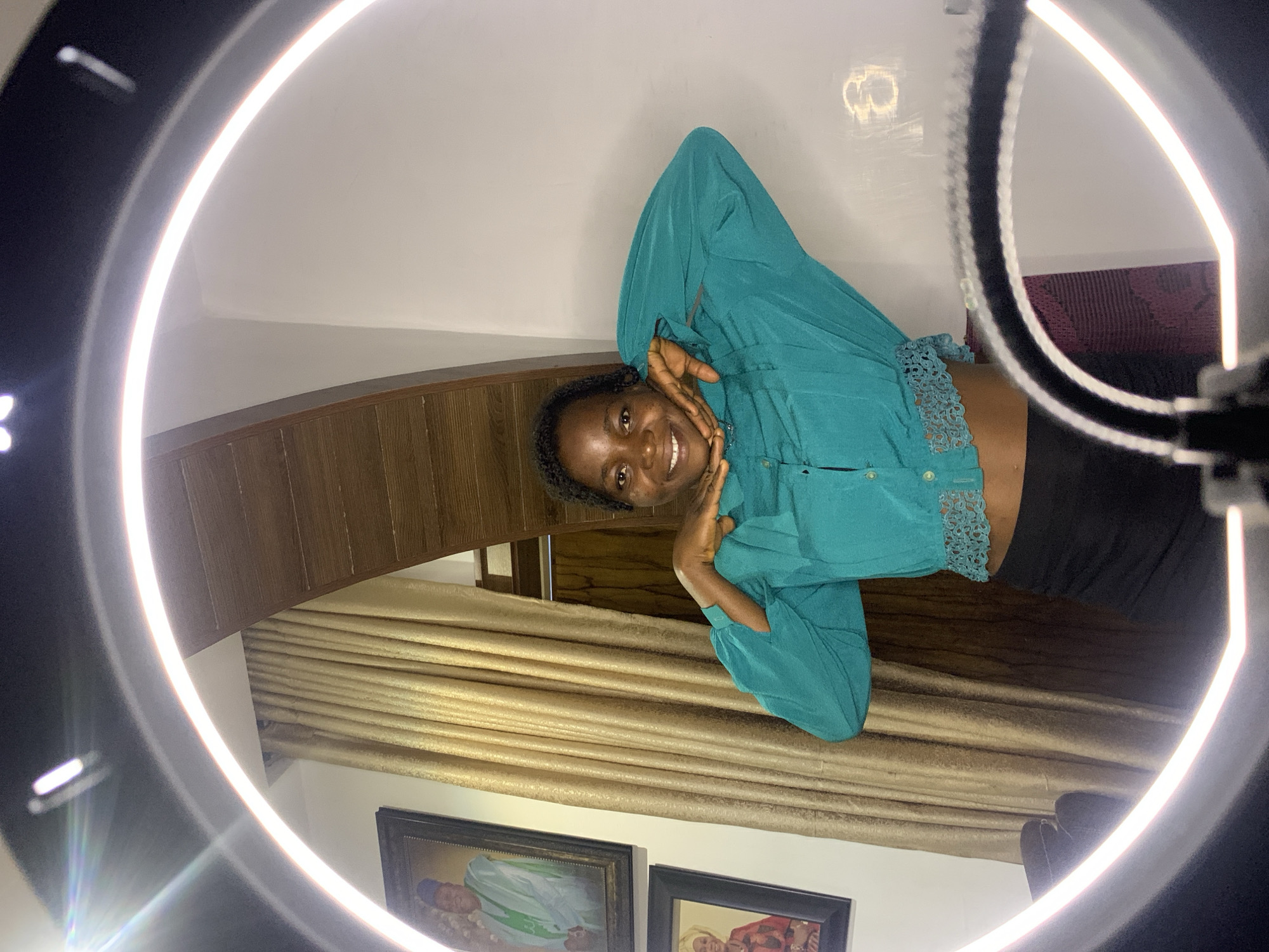 The media file shows a young Black woman posing playfully in front of a ring light. She has dark curly hair and is smiling broadly, with her hands on her cheeks. She is wearing a teal, long-sleeved shirt with a lace detail at the cuffs and a black bottom. The background consists of wooden paneling, gold curtains, and two framed pictures hanging on the wall, one of which appears to depict a religious figure. The overall impression is that of a casual, indoor setting, possibly a home or a studio, with the bright ring light creating a studio-like atmosphere. The lighting suggests the photo was taken indoors, likely during the day or under artificial light. The mood is cheerful and energetic. There is no visible text in the image. The location context is Jos, Nigeria.