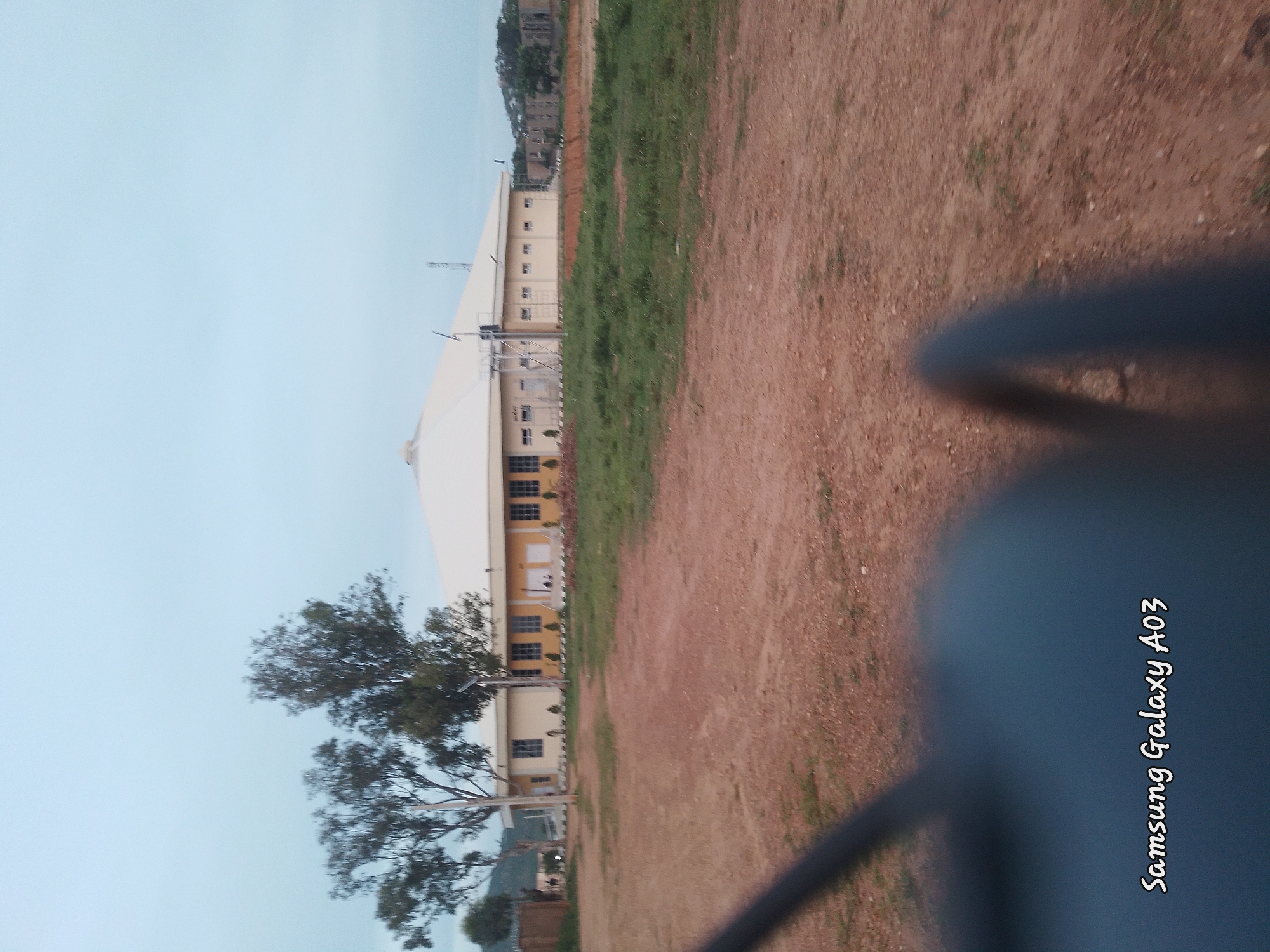 The image displays a wide shot of an outdoor scene, likely taken in Jos, Nigeria. The perspective is from a low angle, looking up towards a large building with a white, tent-like roof, possibly a stadium or a large event hall. The building has a beige-colored lower section with multiple windows. Adjacent to the building is a strip of green grass, and beyond that, a dirt path or field. In the background, there are hills with some structures and trees, suggesting an urban or semi-urban environment. A large tree with dark green foliage is prominent in the foreground, partially obscuring the building. The sky is a clear, pale blue, indicating fair weather and likely daytime, possibly late afternoon or early morning due to the soft lighting and shadow.
The image is oriented vertically, with the "Samsung Galaxy A03" text visible in the bottom right corner, oriented to read upwards. This indicates that the photograph was taken with a Samsung Galaxy A03 phone. There are no people visible in the frame. The overall scene appears to be serene and perhaps a bit desolate, with an emphasis on the architecture and the natural elements of the landscape.