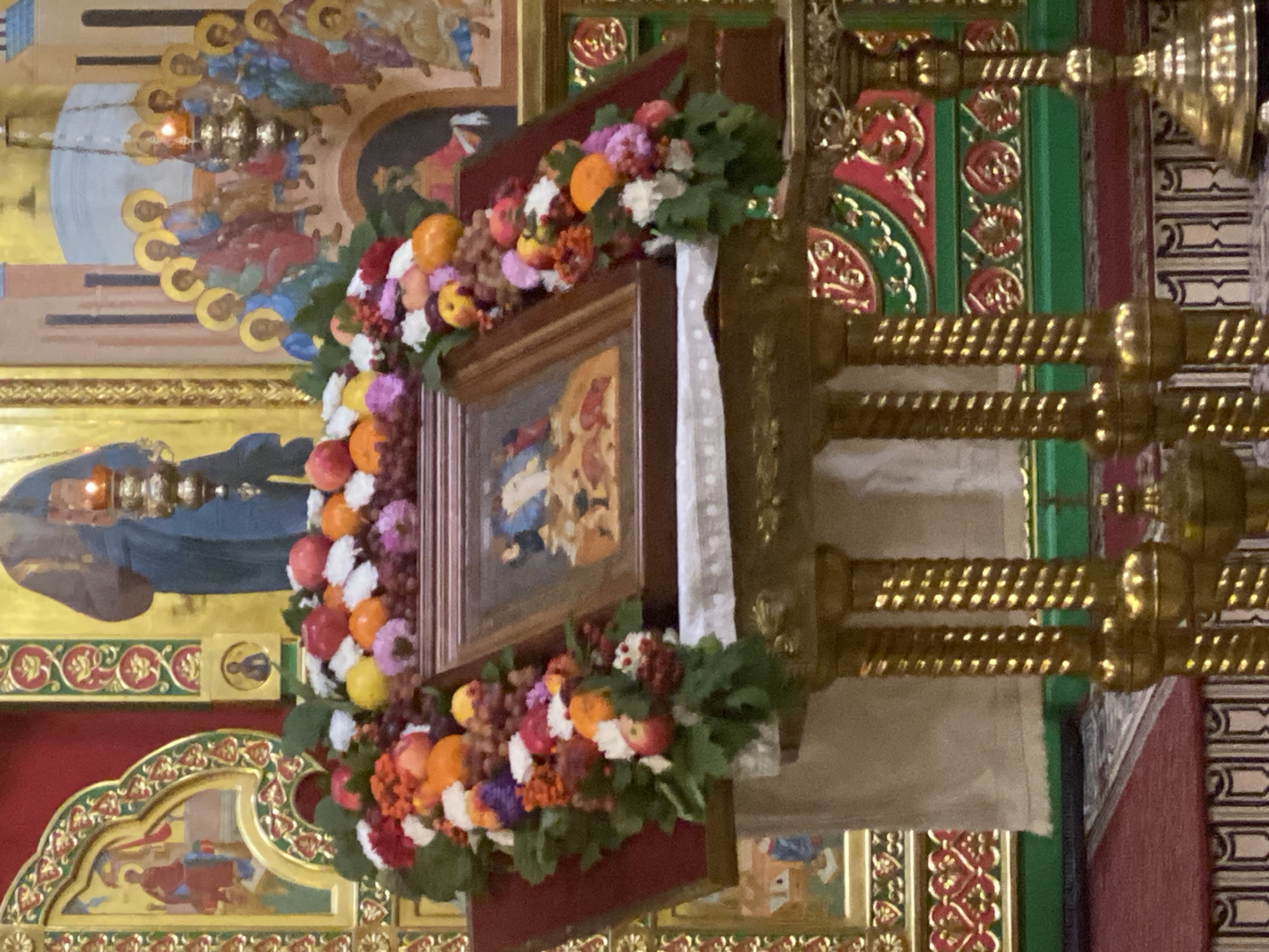 The image shows an iconostasis in an Orthodox church. In the foreground, there is a stand decorated with a garland of fruits, flowers, and berries. On the stand, there is an icon of Mary and Jesus. Behind the icon, an altar with candles and icons can be seen. The overall scene is ornate and religious. Based on visual cues, this appears to be an interior of an Orthodox Christian church, likely in a city like Oktyabrskiy, Russia, given the traditional iconography and style of religious art.