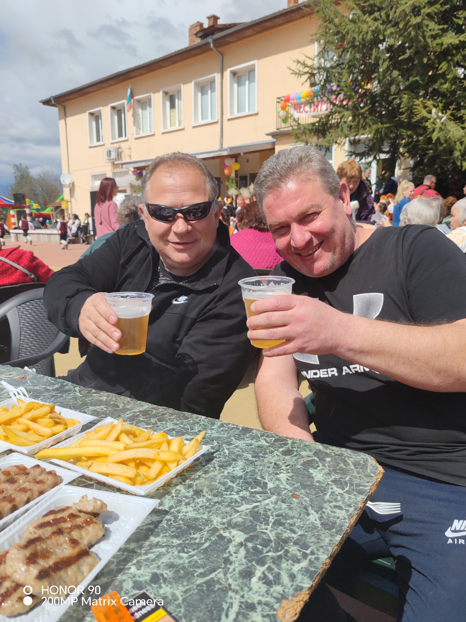 Two men sit at a table outdoors, holding plastic cups filled with a golden liquid. The man on the left wears sunglasses and a black jacket. The man on the right smiles and wears a black Under Armour t-shirt and Nike sweatpants. On the table are two plates of french fries and two plates of grilled patties. The background features a two-story building with a Bulgarian flag and a banner with colorful balloons. A crowd of people is visible behind the table and building. The setting is a public area, likely a festival or celebration, in the city of Rosen, Bulgaria.