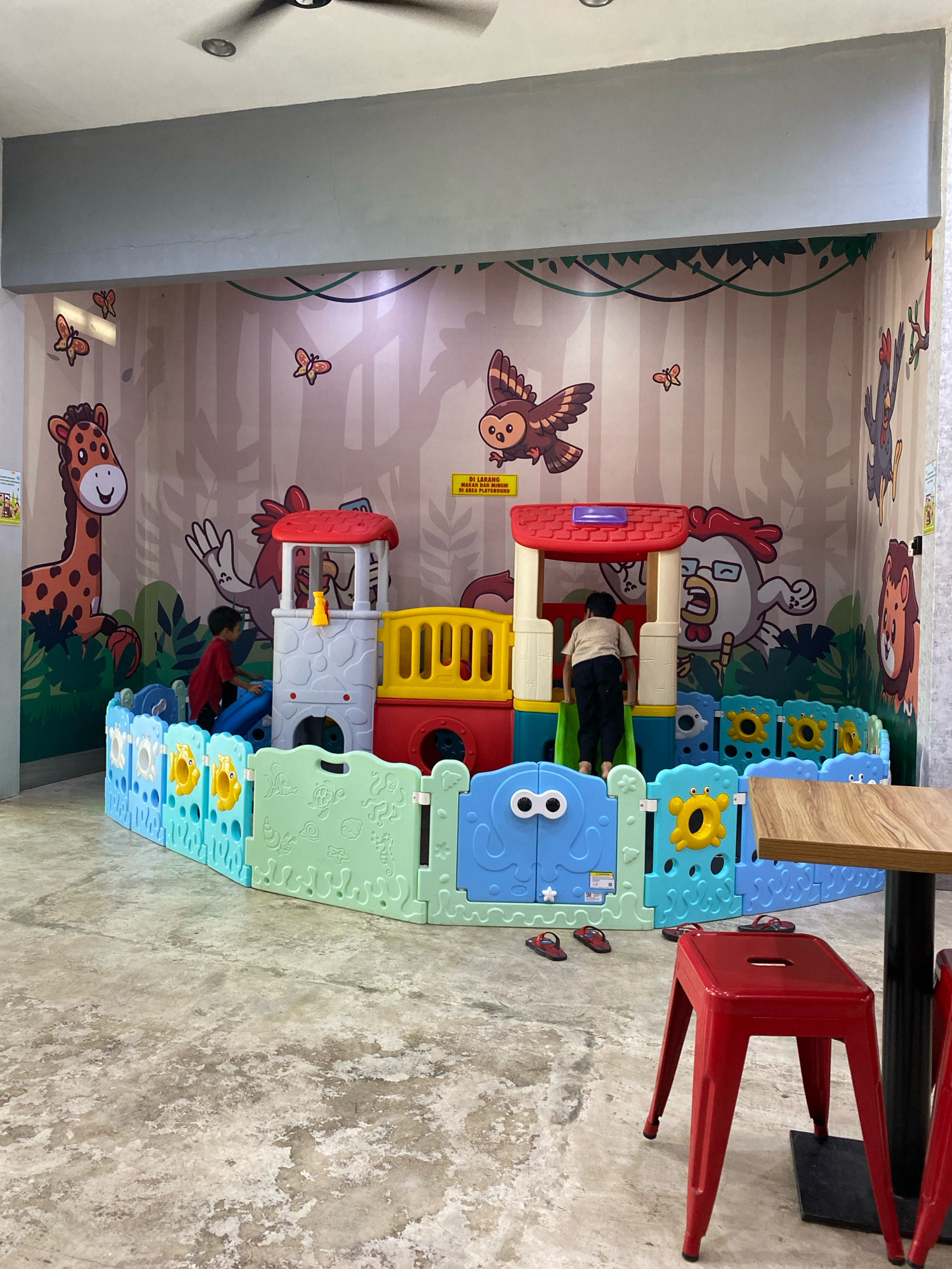The image shows an indoor children's playground in Banjarbaru, Indonesia, with a jungle-themed mural on the wall featuring animals such as a giraffe, owl, chicken, and lion. A colorful plastic play structure is set up within a protective fence. Two young boys are present; one is wearing a red shirt and is near a slide, while the other, in a beige shirt and dark pants, is climbing on the play structure. Several pairs of red flip-flops are scattered on the concrete floor around the play area, and a red metal stool is positioned next to a wooden table. The overall environment appears to be a family-friendly establishment.