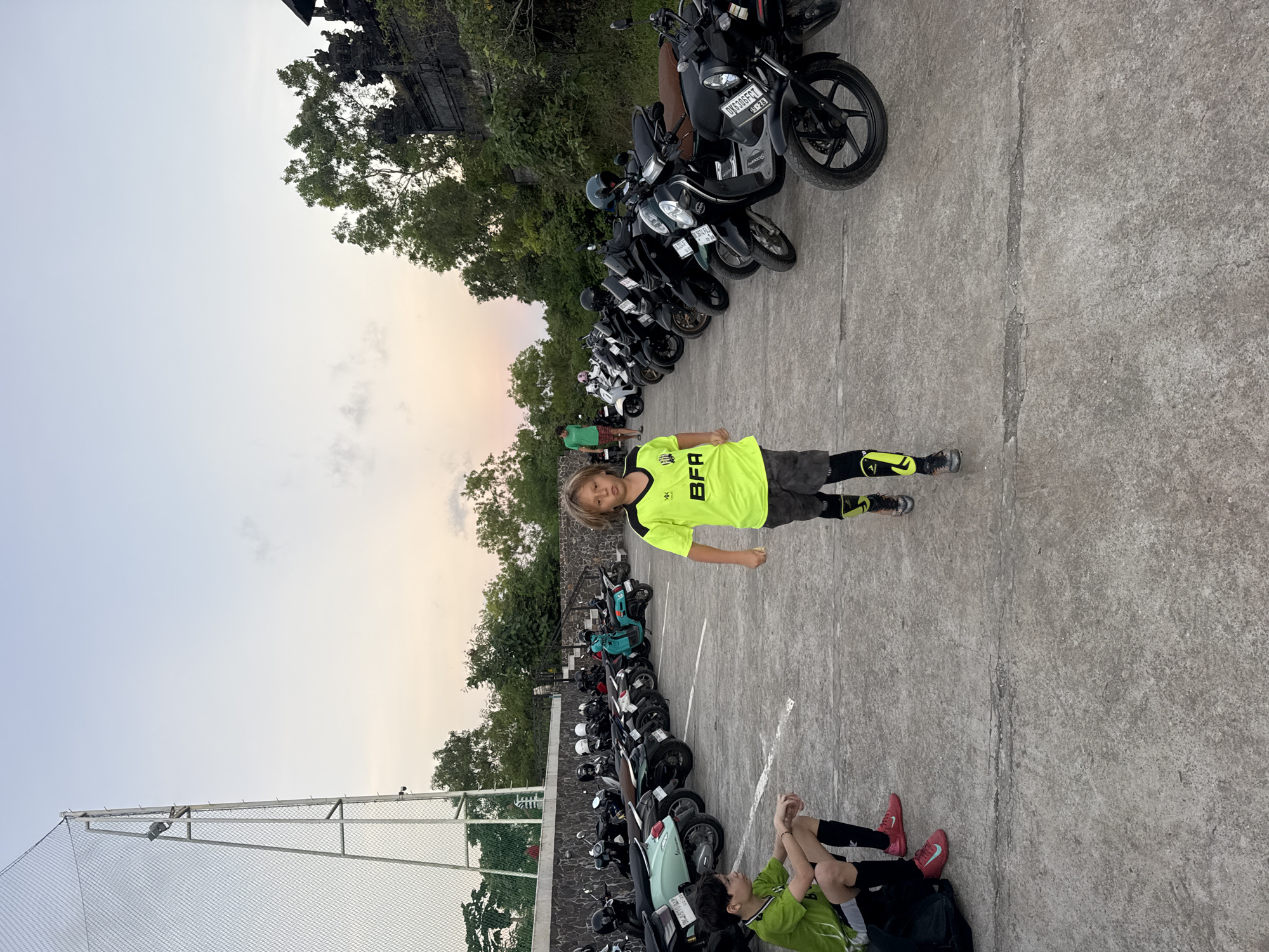 A young Caucasian boy in a neon green soccer jersey and gray shorts stands on a gray concrete lot adjacent to rows of parked motorbikes. Another boy, also in soccer attire, sits on the ground nearby. The background includes greenery and glimpses of a structure that may be related to the nearby football field. The location is in Kuta Selatan, Indonesia.