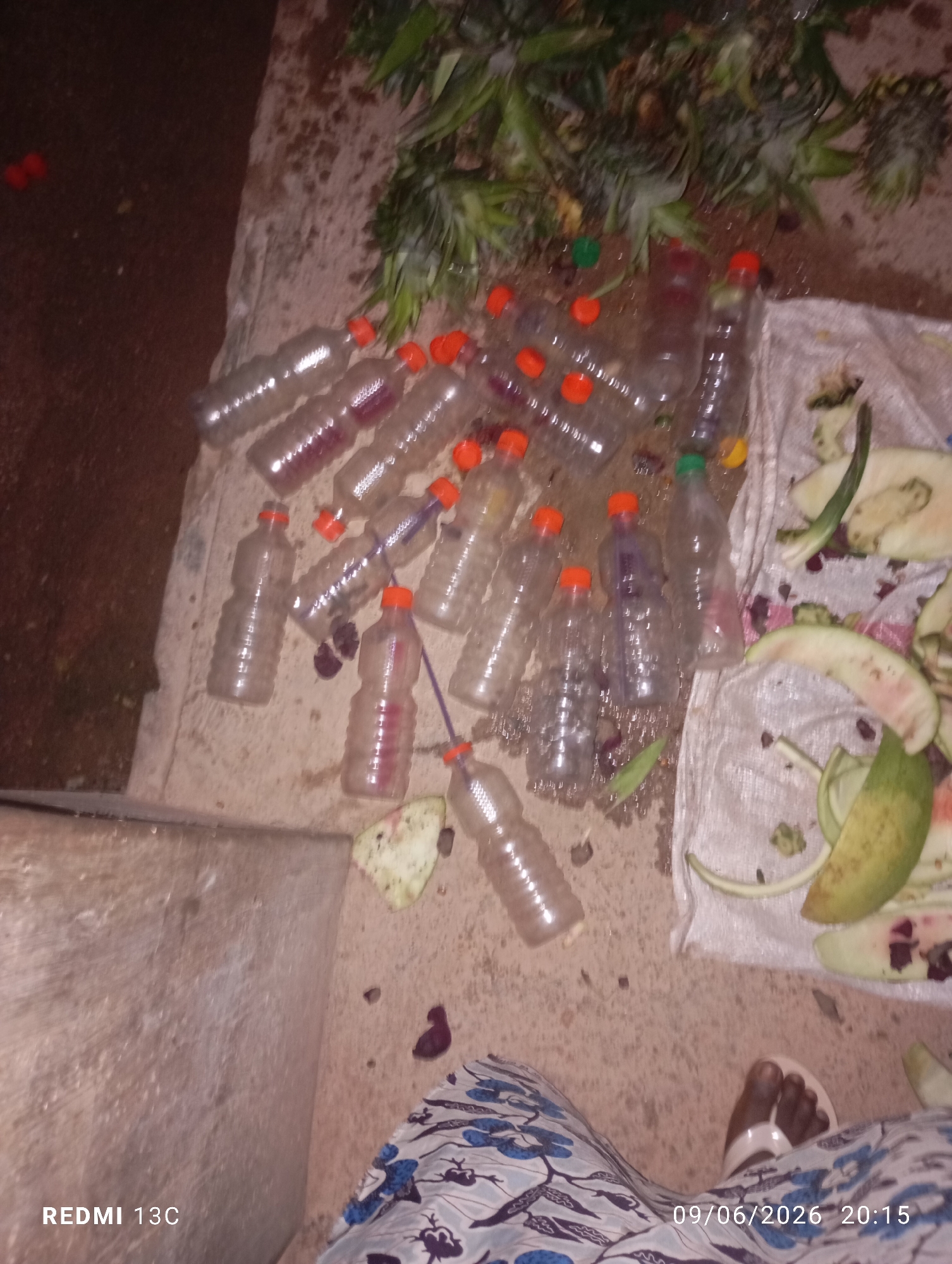 An overhead, low-angle medium shot captures an outdoor scene with various objects scattered on the ground. In the foreground, a person's feet are visible, wearing white sandals, positioned on a patterned cloth. To the left, the edge of a concrete structure is present. A large number of clear plastic bottles with orange caps are distributed across the ground, some containing a reddish liquid. Some bottles are upright, while others are lying on their sides. Alongside the bottles are discarded pieces of fruit, including watermelon rinds, and some green vegetation that appears to be pineapple leaves. The ground itself is a mixture of dirt and concrete. A white plastic sheet is visible in the midground, serving as a surface for some of the fruit waste. The image is dark, suggesting it was taken at night or in low light conditions.