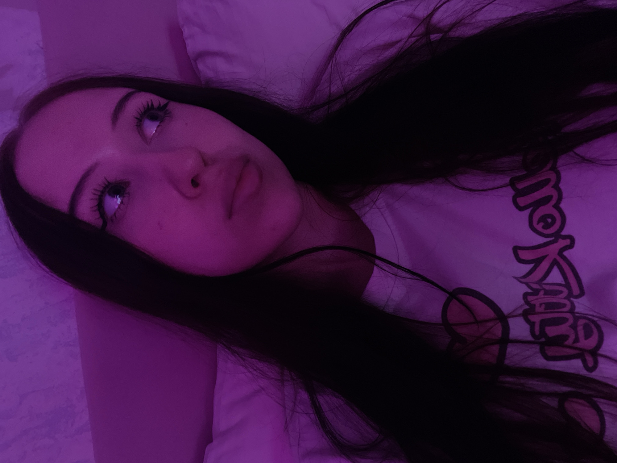 The image shows a close-up portrait of a young woman with long, dark hair, illuminated by pinkish-purple light. She is wearing a white t-shirt with a graphic design, which appears to be pink text with a black outline and a pink heart symbol. The woman is looking away from the camera to her right, with her eyes slightly widened and her mouth slightly open. She has long, dark eyelashes and appears to be lying down, possibly on a bed, as indicated by the soft, pillow-like texture in the background. The environment is dimly lit and the dominant color is a vibrant pinkish-purple, suggesting the use of colored lighting. There is no discernible information within the image to indicate a specific location in Votkinsk, Russia.