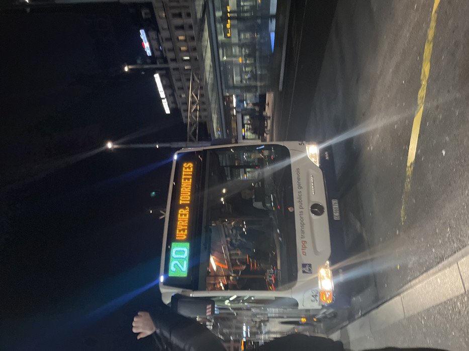 This night-time image, taken in Genève, Switzerland, shows a white city bus stopped at what appears to be a bus stop or street-side location. The bus, possibly a Mercedes-Benz (indicated by the logo), has its headlights on, illuminating the immediate area. Its electronic destination display clearly shows "20" in green for the route number and "VEYRIER, TOURNETTES" in amber for the destination.
The side of the bus features the text "tpg transports publics genevois", identifying it as part of Geneva's public transport, along with a wheelchair accessibility symbol. A unit number "543" is visible on the front, and the license plate "GE-900763" further confirms the location as Geneva.
In the foreground, the arm and shoulder of a person wearing a dark jacket are visible. The background includes several illuminated buildings, suggesting an urban environment. A distinctive red illuminated sign, likely "Swisscom", is visible on one building, along with other less discernible white illuminated signs. Streetlights punctuate the dark sky, casting rays of light. The ground is dark asphalt with a yellow painted line. The scene depicts a quiet moment in a city at night.