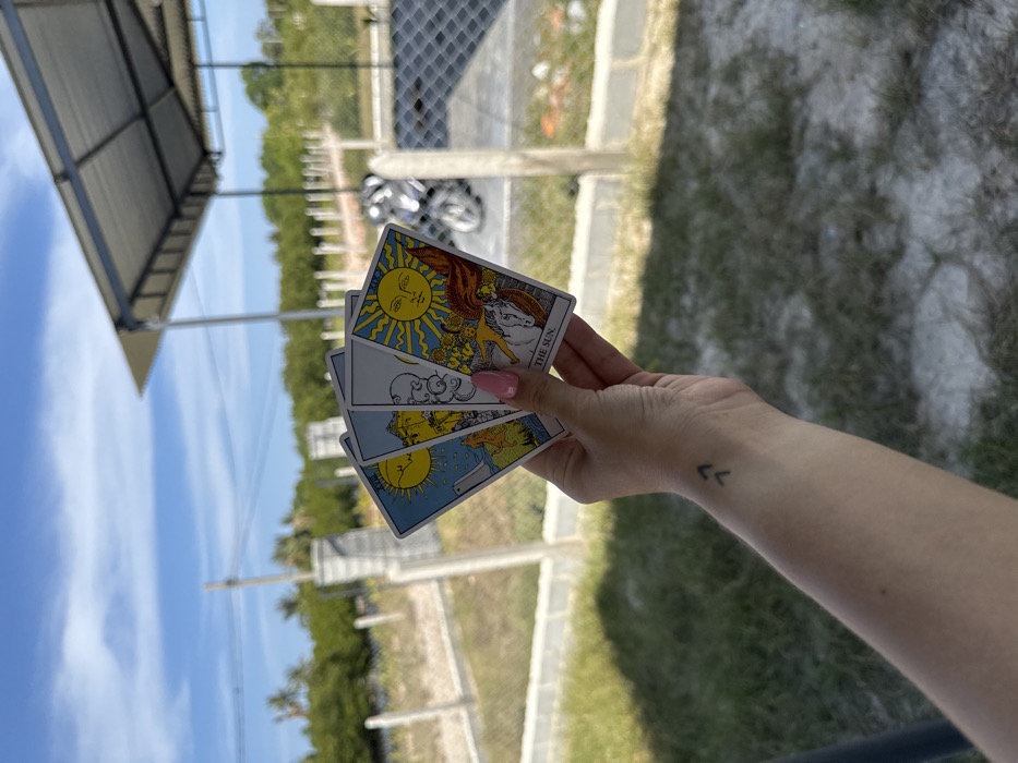 A light-skinned human hand, with pink manicured fingernails, holds four fanned-out tarot cards against an outdoor backdrop. A small black tattoo of two upward-pointing chevron symbols is visible on the inner wrist. The prominent rightmost card is "The Sun" from a Rider-Waite-style deck, depicting a large yellow sun, a naked child on a white horse, and sunflowers. The other three cards, partially obscured, also feature celestial or symbolic imagery.
The scene is set outdoors in Cam Đức, Vietnam, under a clear blue sky with sparse white clouds. The foreground consists of dry, patchy green grass and light-colored soil. A fence line, composed of light-colored concrete posts and horizontal wire strands, separates the immediate foreground from the mid-ground. Beyond this fence, to the right, a white motorbike is partially visible. A modern structure with a light-colored corrugated metal roof supported by dark poles is present in the mid-ground. The far background features a dense line of green foliage or trees, with utility poles and overhead wires visible against the sky.