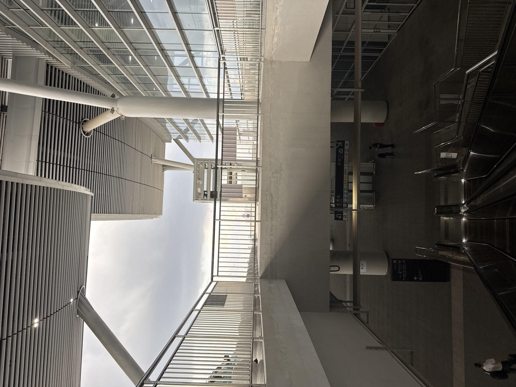 The image depicts a modern transportation hub, likely a train station or airport terminal, in Huizhou, China. The architecture is characterized by large glass windows, metal railings, and a covered walkway with a sloped ceiling. Natural light filters through the overcast sky, illuminating the clean lines of the structure. In the foreground, escalators lead down to a lower level, where a few individuals are present. One person in a white shirt and dark pants is walking away from the camera with a backpack. Another individual, dressed in dark clothing, stands with a small luggage cart. Signage with Chinese characters is visible, indicating directions and facilities. The environment appears to be orderly and functional, designed for efficient passenger flow.