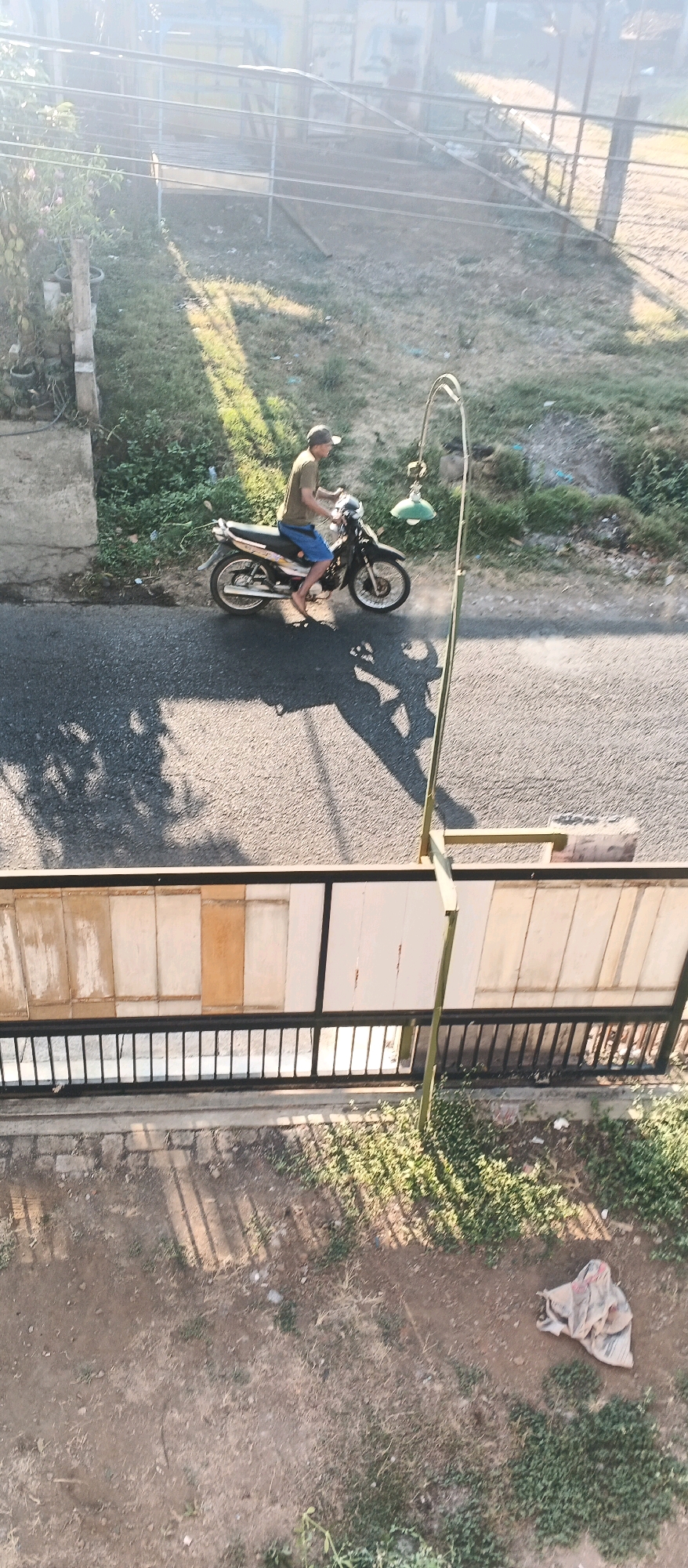 A person wearing a cap, a brown t-shirt, and blue shorts rides a black motorcycle with a yellow stripe on a paved road. The motorcycle has a white panel on its side with some writing. A green street lamp with a white shade hangs from a pole to the right of the motorcycle. The road is bordered by a grassy area with some bushes and a white wall with horizontal planks. A black metal fence runs along the bottom of the image, in front of another wall with vertical planks. The ground beyond the fence is covered in dirt and sparse green vegetation, with a bundle of cloth lying on the ground. The scene appears to be outdoors, possibly in Bocek, Indonesia, given the context. The sun is casting shadows on the road and ground.