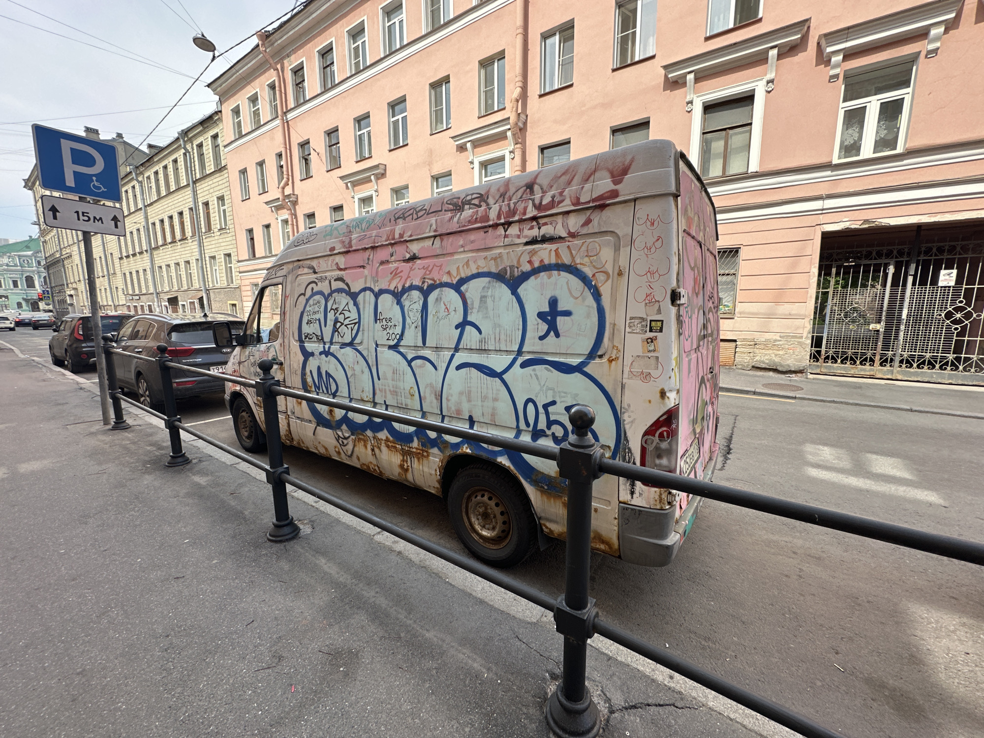 The image depicts a graffiti-covered, rusty white van parked along a street in Saint Petersburg, Russia. A black metal railing separates the sidewalk from the road. Behind the van, a parking sign with a wheelchair symbol indicates accessible parking within 15 meters. Several parked cars are visible further down the street. The background features buildings with a mix of light pink and yellow facades. The sky is overcast.