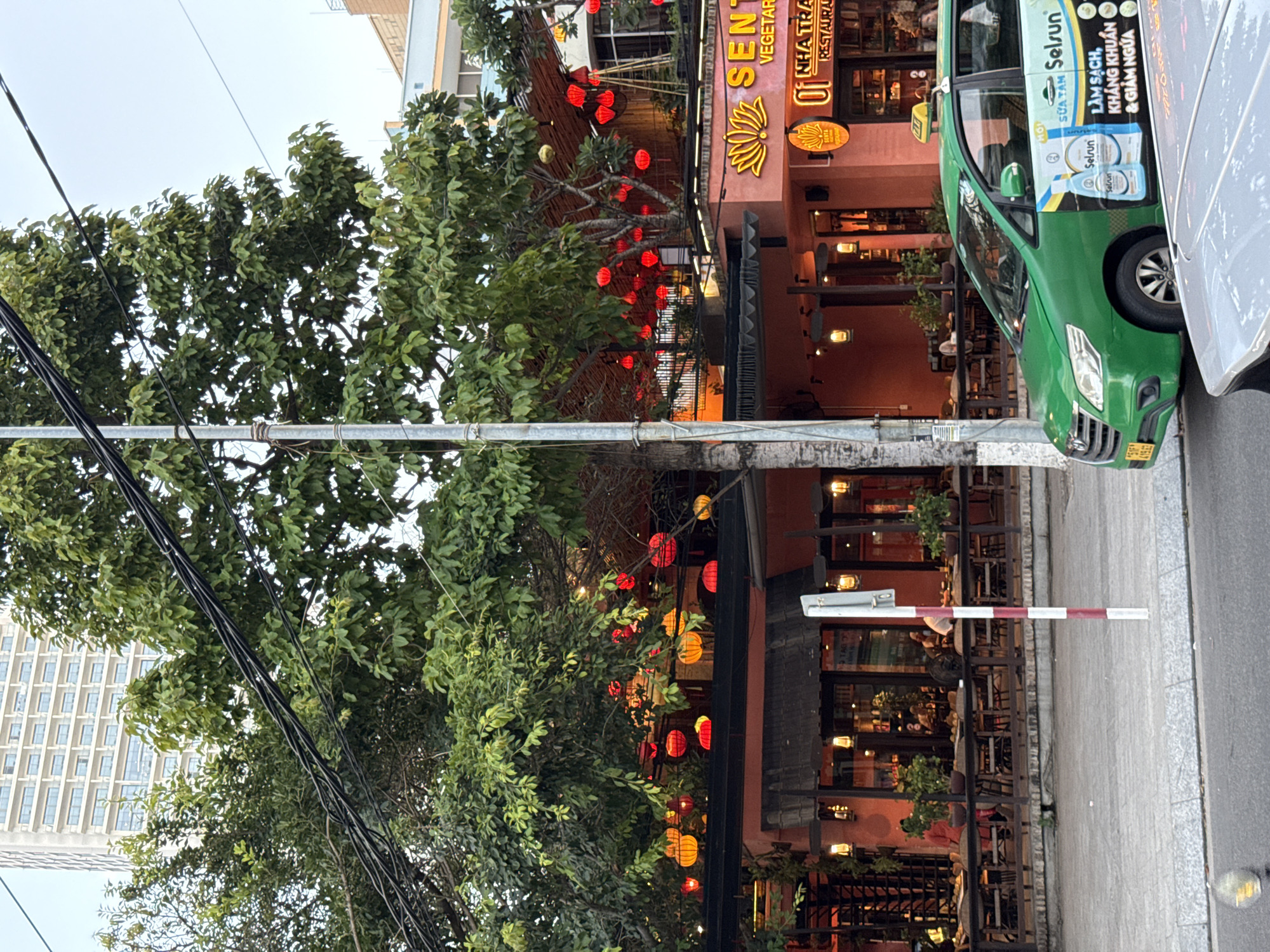 The image depicts a street scene in Nha Trang, Vietnam, featuring a vibrant restaurant with red lanterns strung across its facade and through the surrounding trees. The restaurant, identified by signage as "SENTOSA VEGETARIAN RESTAURANT 01 NHA TRANG RESTAURANT," has an outdoor seating area with tables and chairs visible. A green taxi is parked in front of the restaurant, partially obscuring the lower portion of the building. The background shows a tall, modern building, suggesting an urban environment. A street pole with red and white markings stands on the sidewalk. The lighting suggests it is either late afternoon or early evening.
