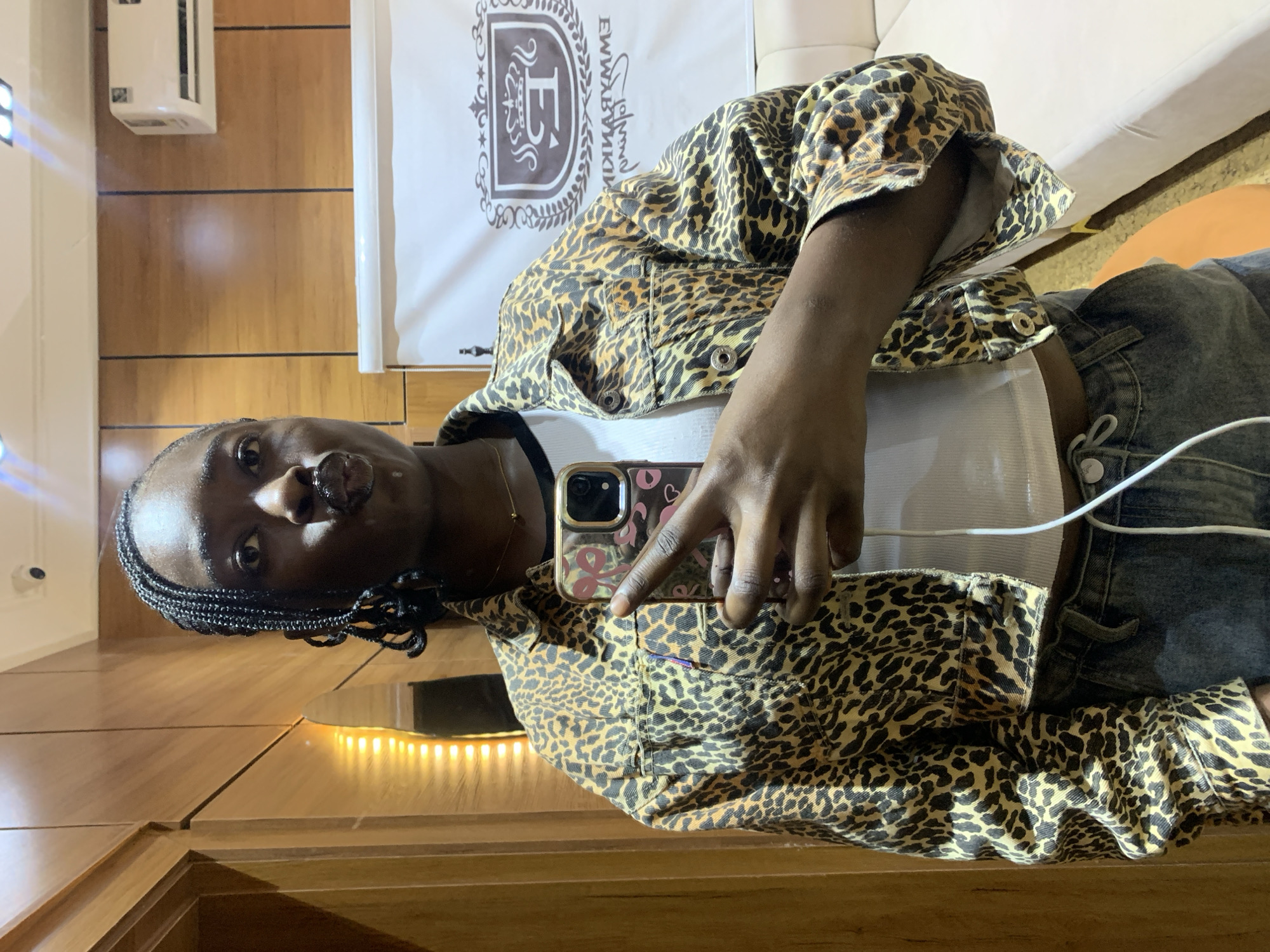 A woman with dark skin and braided hair is taking a selfie. She is wearing a leopard print jacket over a white crop top and blue jeans. She has her lips pursed, as if for a kiss. Her right hand is holding a phone, which displays a sticker of a pink bow and a pink heart. The background appears to be an indoor space with wood-paneled walls and a white banner with a crest. A white air conditioning unit is mounted on the wall. The lighting suggests it is daytime, but the scene could be indoors during any time of day. It's not possible to determine the exact location in Gura Top, Nigeria from the image.