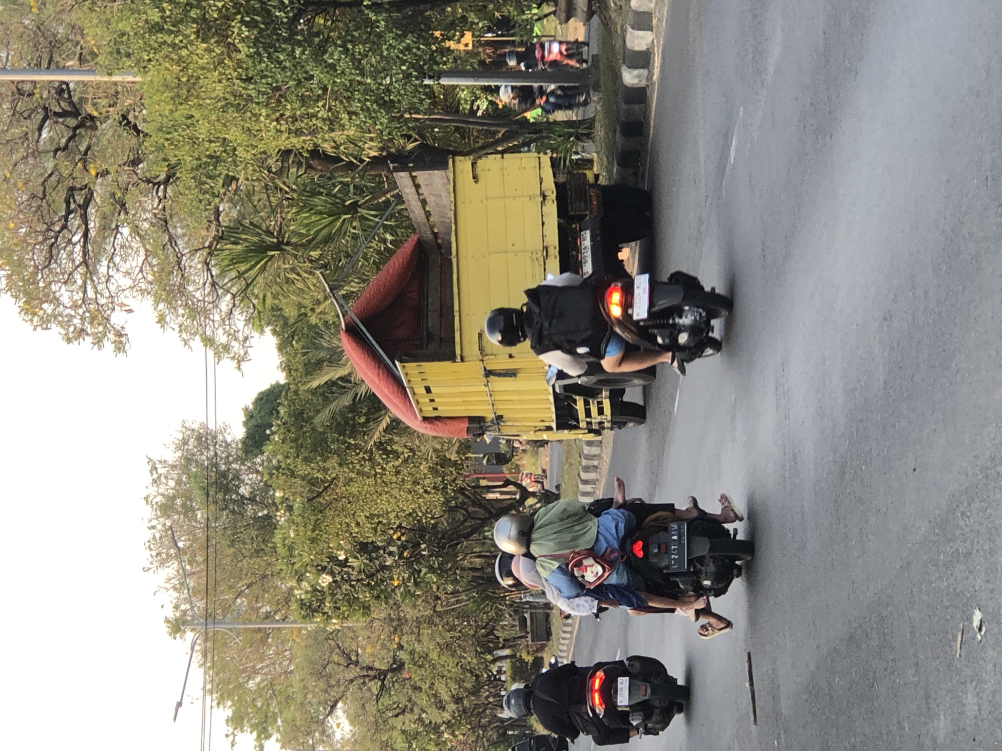 In Ngaban, Indonesia, a yellow truck carrying a large, red tarp is driving down a paved road. Several motorcycles are on the road as well, some with multiple passengers. The motorcycles are in motion, and the people on them are wearing helmets and casual clothing. The surroundings include lush green trees and vegetation, with a bright sky overhead. The scene appears to be a typical street view in a tropical urban or suburban area.