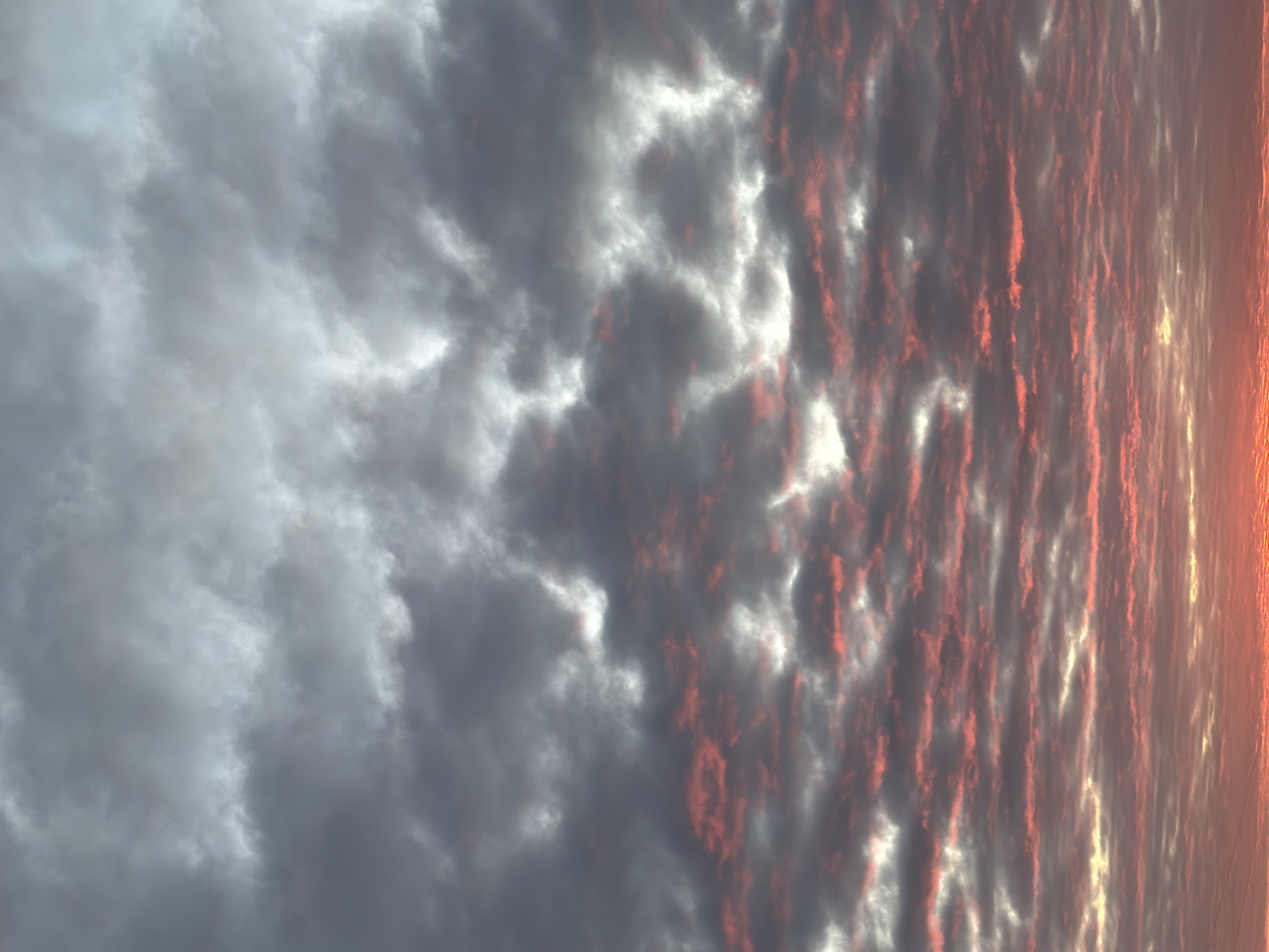 This image depicts a wide vertical view of a sky over Chaikovsky, Russia, during what appears to be either sunrise or sunset. The upper portion of the sky is dominated by dense, layered grey and white clouds. Below this, a transition occurs where dark grey clouds are illuminated from beneath, revealing numerous elongated and patchy formations highlighted with vibrant red and pink hues. Towards the bottom of the frame, the sky transitions into a bright orange and reddish glow, indicating the low position of the sun below the visible horizon. The cloud formations vary from soft, diffuse masses in the upper section to more textured, wave-like patterns and streaky illuminated patches in the lower two-thirds of the image. No ground features, structures, people, or discernible objects other than the sky and clouds are visible.