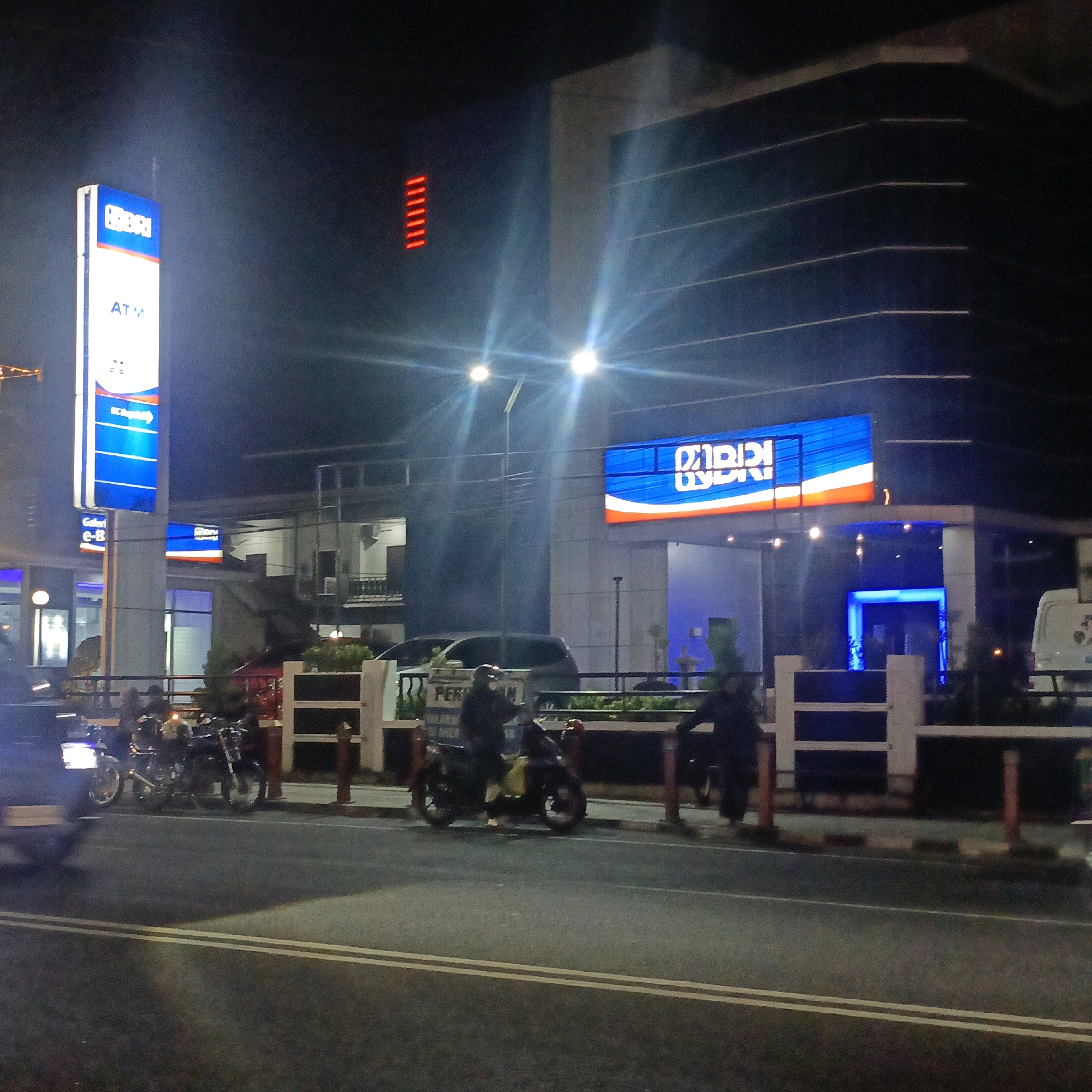 In Boyolali, Indonesia, at night, the scene depicts a street in front of a BRI (Bank Rakyat Indonesia) branch. A tall, illuminated sign for BRI and ATM is prominently displayed on the left. Adjacent to it, another BRI sign is visible. The building's facade is modern with horizontal lines and bright lights. Several motorcycles are parked or being ridden on the street, with some people visible on them. The street itself is asphalt with double yellow lines. A white van is parked on the right side of the image. The overall atmosphere is that of a well-lit urban environment at night.