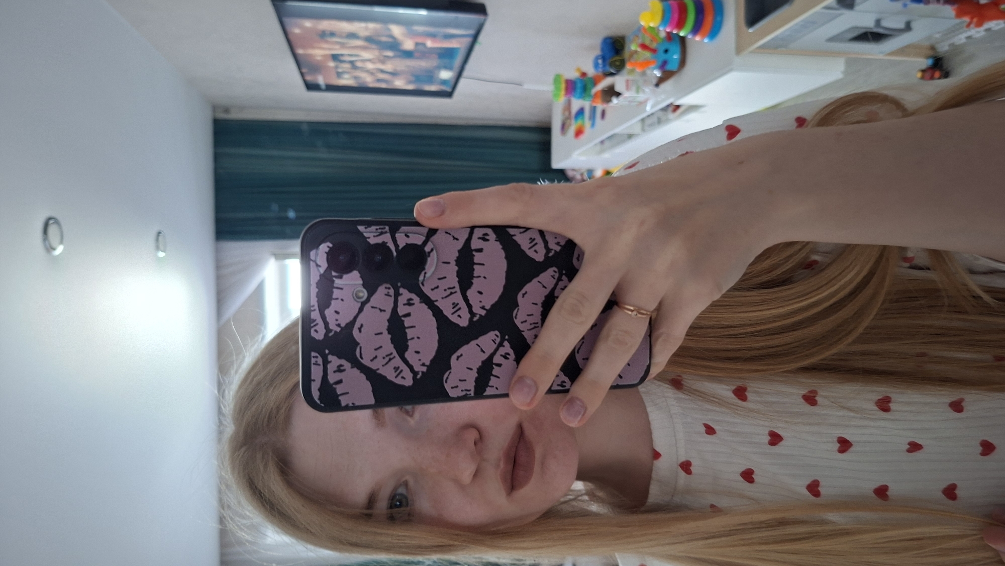 A blonde woman takes a selfie with a black smartphone with a light purple lips design. She's indoors. A television is mounted on the wall behind her, and a shelf filled with toys is visible to the right. The room is located in Chaikovsky, Russia. The wall is white with two circular attachments. She is wearing a white shirt with small red hearts.