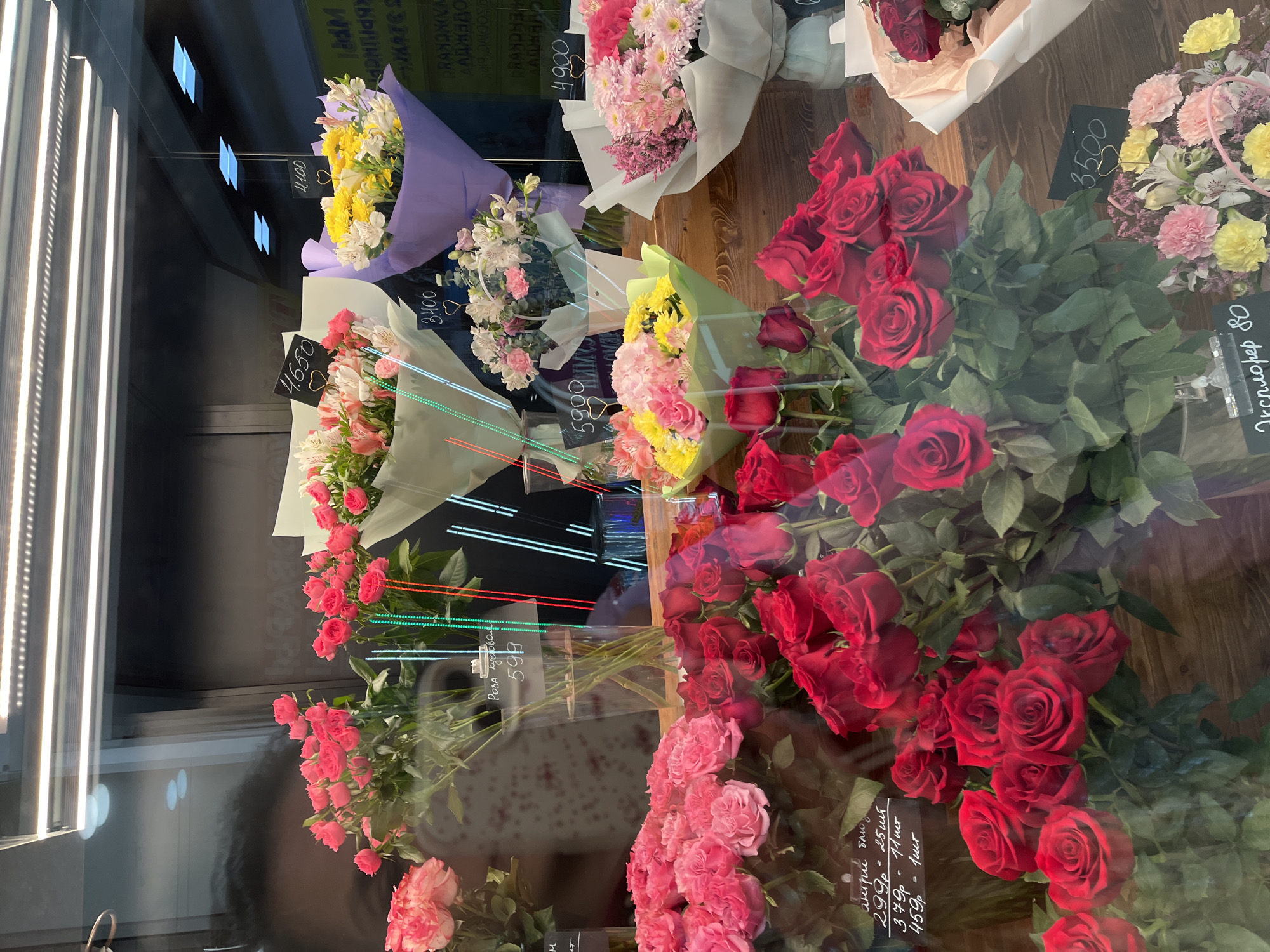 The image shows a florist's shop display in Petropavlovsk-Kamchatsky, Russia. There are numerous bouquets of flowers including red and pink roses, yellow and white alstroemeria, and mixed flower arrangements. Price tags are visible for some bouquets, displaying prices in rubles. A reflection of the shop's interior, including the display lights and possibly a person, is visible in the glass. The arrangement of the flowers suggests they are for sale in a flower shop.