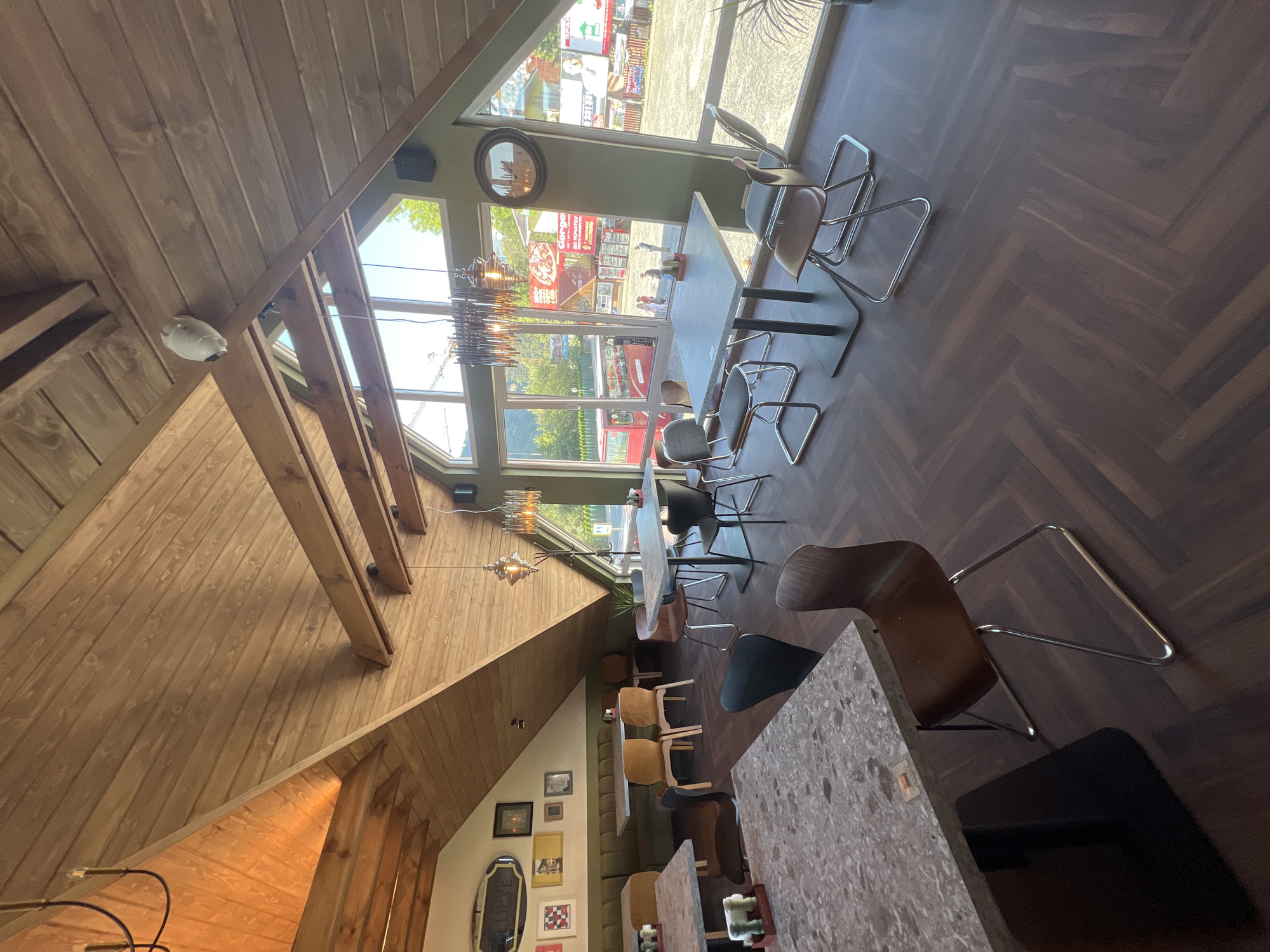 The image depicts the interior of a restaurant in Yaremche, Ukraine, featuring a sloped wooden ceiling and large windows offering a view of the street. Several tables with modern chairs are arranged throughout the dining area. The flooring is a dark wood parquet. One table in the foreground has a light-colored speckled surface and is set with condiments. The walls are painted a muted green and are adorned with framed artwork. A long bench seat is visible along the back wall. The lighting fixtures are contemporary. Outside, a red double-decker bus and various signs are visible.