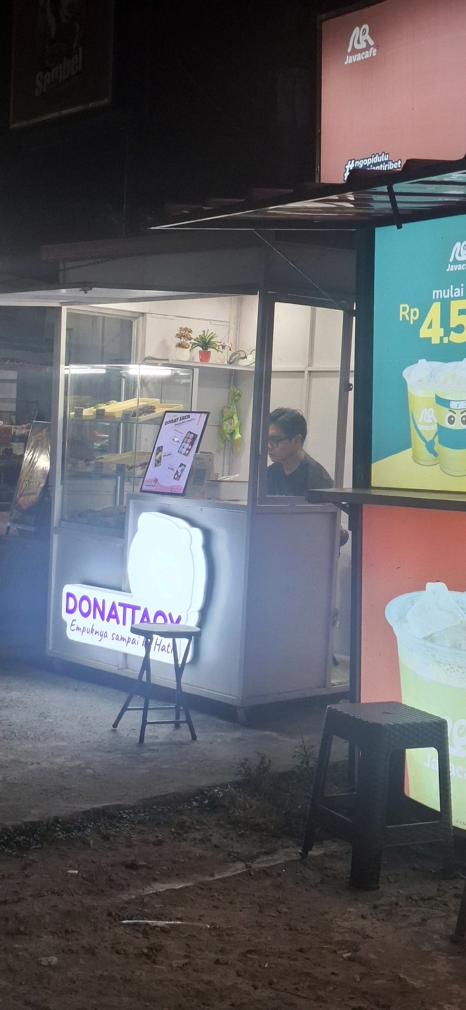 The image shows a street-level view of a small food stall at night in Pabelan, Indonesia. The stall is illuminated by internal lighting and external signage. A neon sign on the left side of the stall reads "DONATTACK" in purple lettering, with smaller text below it that appears to be in Indonesian. Inside the stall, donuts are displayed in a glass case. A person wearing glasses and a dark t-shirt is visible behind the counter. To the right of the stall, a large advertisement for "Javacafe" is displayed on a screen, showing a price of "Rp 45." Several plastic stools are placed outside the stall. The ground in front of the stall is paved and appears wet.