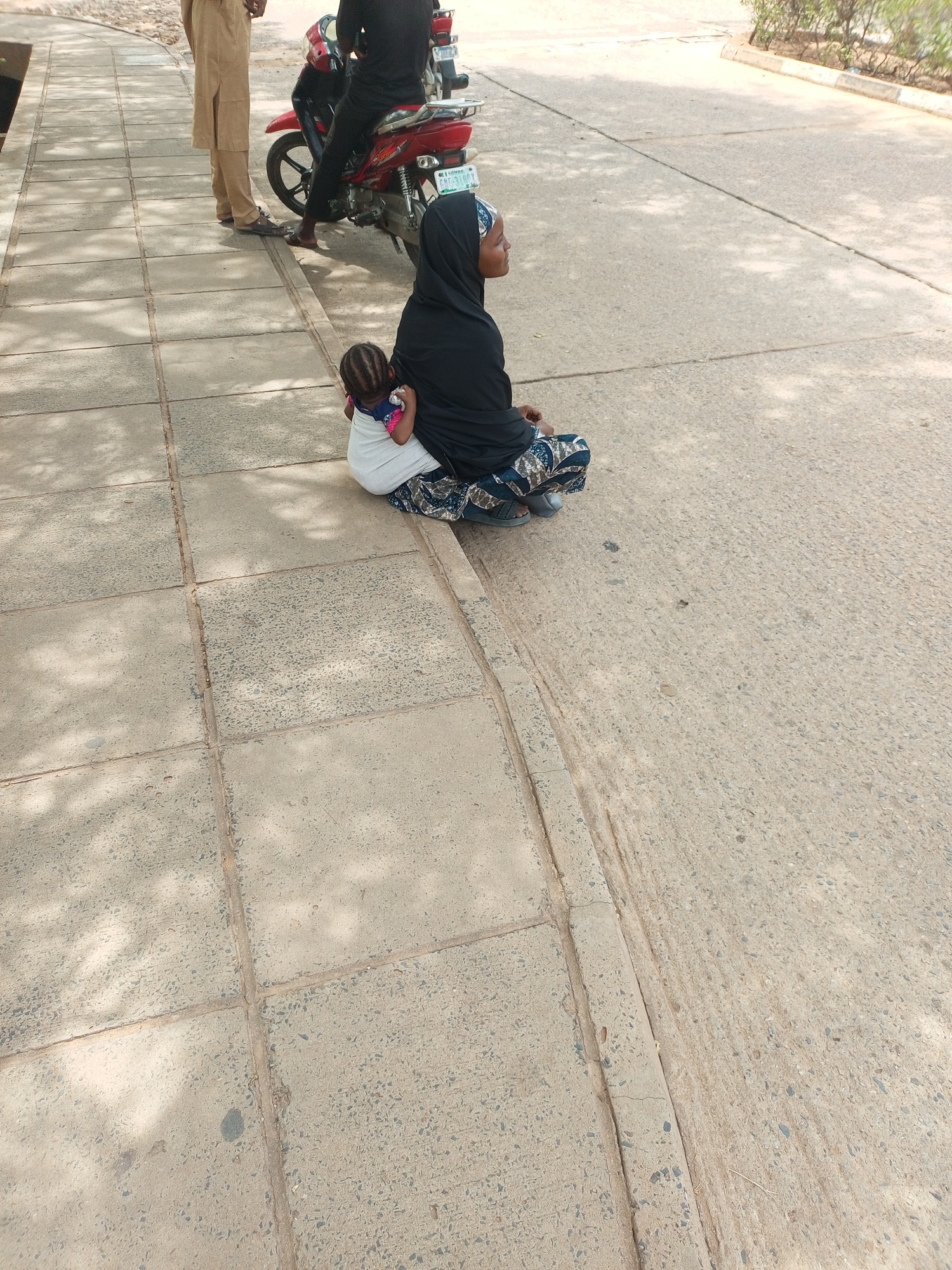 A woman in a black hijab sits on a sidewalk near a street in Gombe, Nigeria, holding a child on her back. Two men stand near a red motorbike behind her. The setting appears to be a public area with paved surfaces and some vegetation in the background.