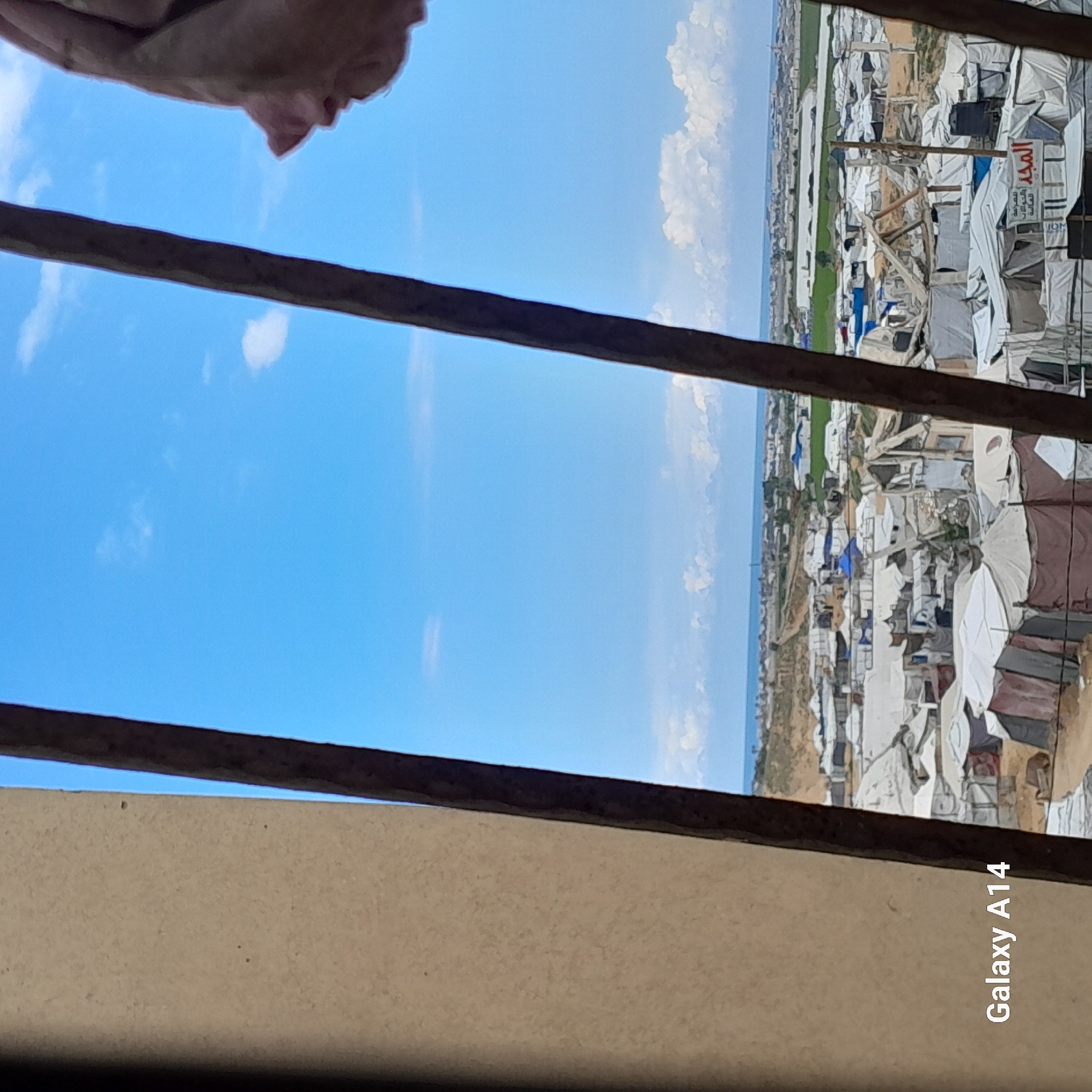 The image shows a view of a refugee camp near a body of water. The camp is densely populated with tents and makeshift structures. The sky is blue with some clouds. Part of a window frame or similar structure is in the foreground. In the top left corner, a portion of a face is visible. The location is Unknown, Unknown.
