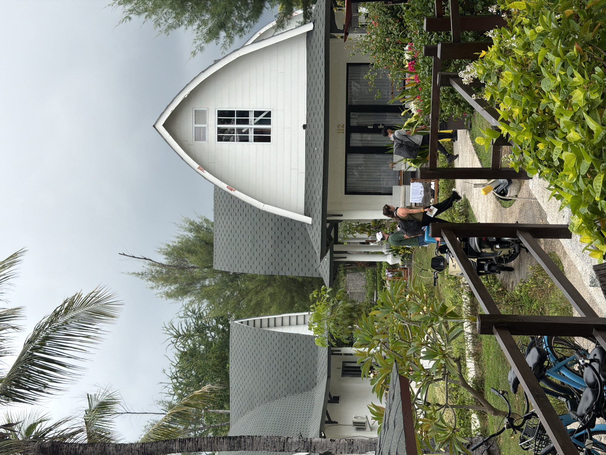 The image shows a resort or hotel scene in an outdoor environment, likely in Indonesia given the palm trees and architecture. In the foreground, a wooden fence separates the viewer from a pathway. On the pathway, two people are walking away from the camera. One person, wearing a green shirt and white cap, walks towards the right. Beside them, a person with dark hair and a backpack also walks towards the right. Further in the background, two houses with distinctive gambrel roofs are visible. The house on the right has the number "112" above a set of glass doors. The houses are white with dark trim and grey shingled roofs. Lush green vegetation and flowering plants surround the buildings. Several bicycles and a scooter are parked near the pathway. The sky is overcast.