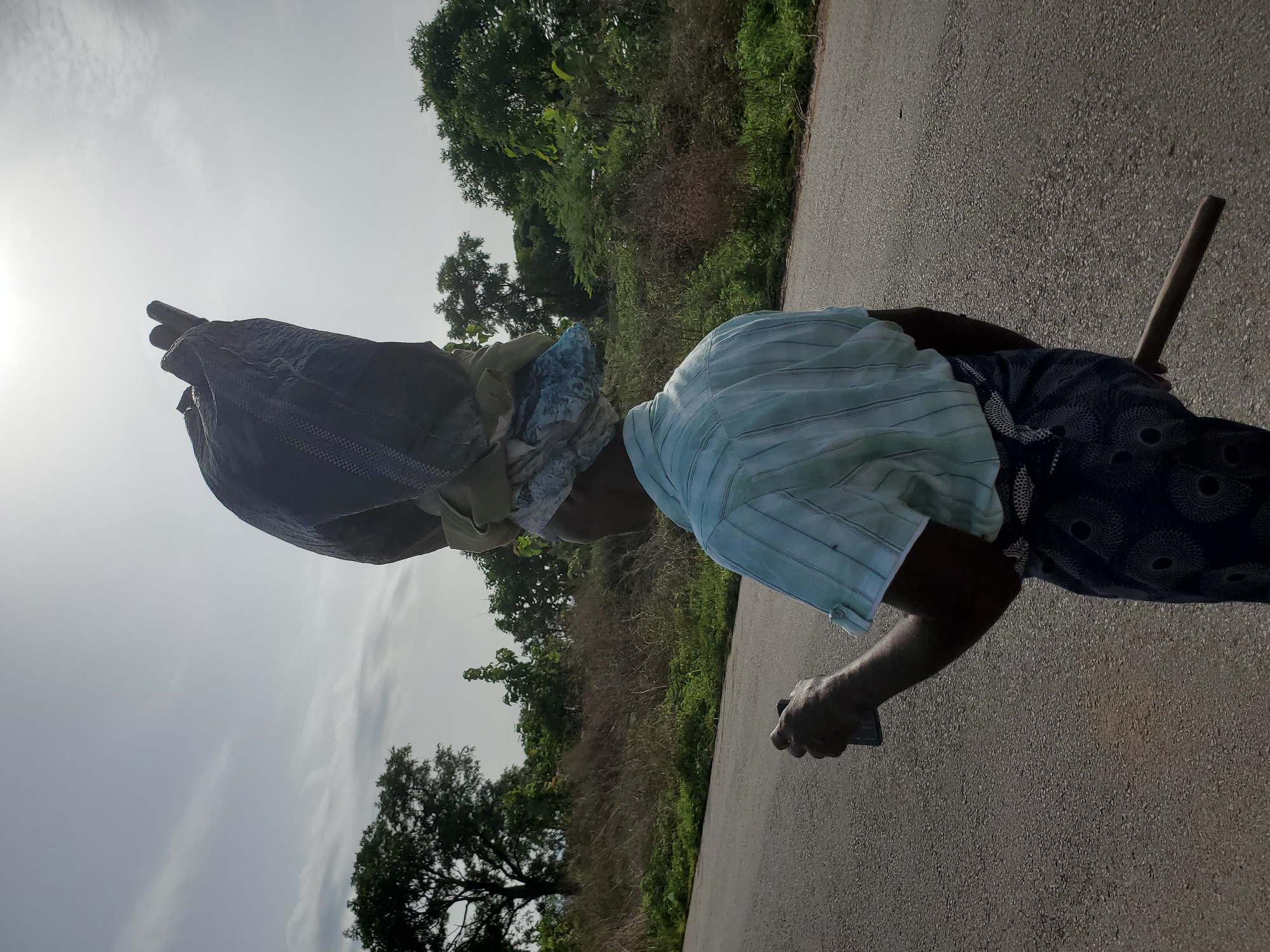 A person is walking along a paved road, likely in a rural area, based on the surrounding vegetation, on the outskirts of the city of Mararaba, Nigeria. The person is carrying a large bag or load on their head, secured with cloth wrappings. They are also holding a stick in one hand. The person is wearing a light-colored, striped shirt and a patterned, dark-colored bottom. The sky is bright, suggesting it is daytime. The scene includes roadside vegetation, trees, and a paved surface. The person is the primary subject and appears to be traveling on foot. The location is inferred as being on the edge of the city of Mararaba based on typical land use patterns.