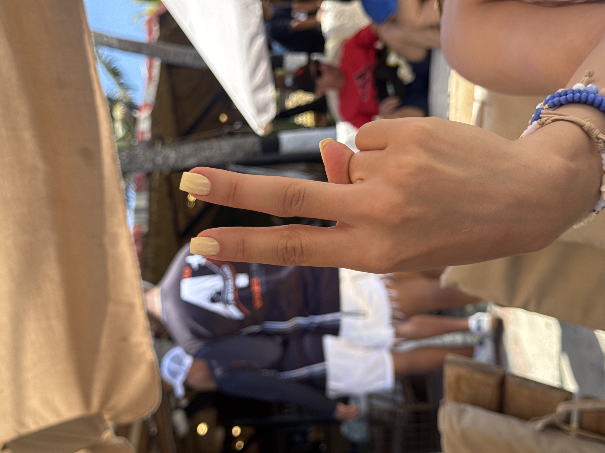 A close-up, slightly rotated shot depicts a hand displaying two fingers in front of a blurry background. The hand has pale yellow nail polish. A bracelet adorns the wrist. In the background, people are seen standing around tables underneath a covered structure. The scene is possibly outdoor given the lighting. The background is blurry. The image does not clearly show Porlamar or Venezuela.