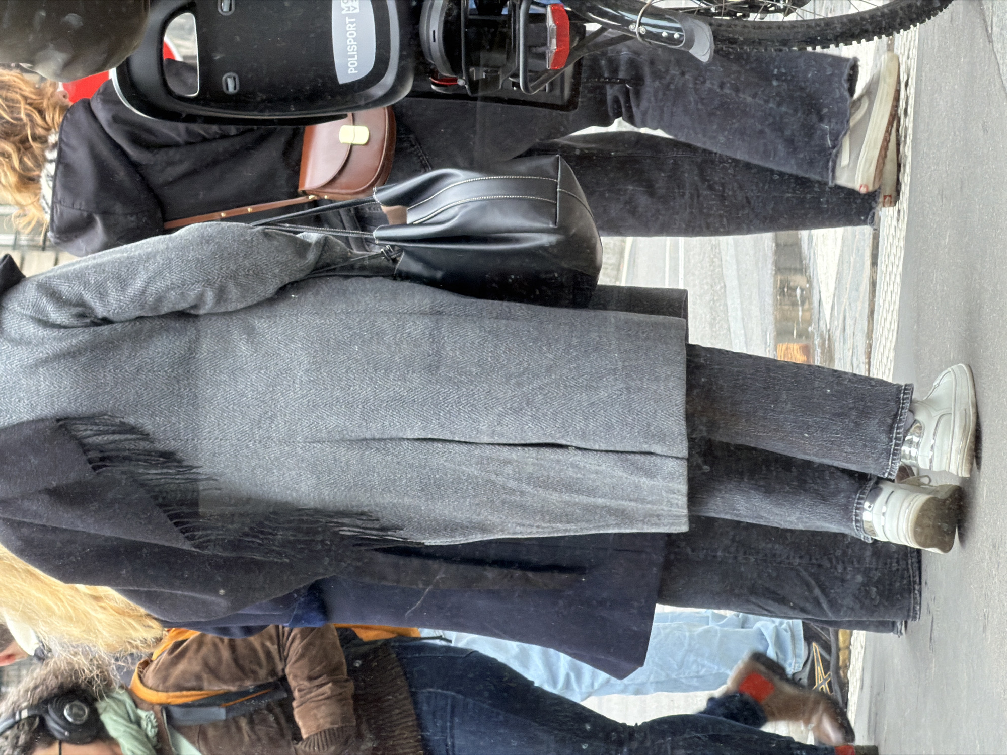 The image appears to have been taken in an urban setting, likely in Paris, France, judging by the architecture and street features visible. A person wearing a long gray coat, dark jeans, and white sneakers stands on a sidewalk. The person carries a black shoulder bag and is positioned next to a bicycle equipped with a child carrier seat. Another individual, wearing a brown jacket and blue jeans, is also present, though their full figure is not clearly visible. The setting is a typical Parisian street, with light-colored pavement and possible reflections indicating wet conditions. The overall scene suggests an everyday moment in the city.